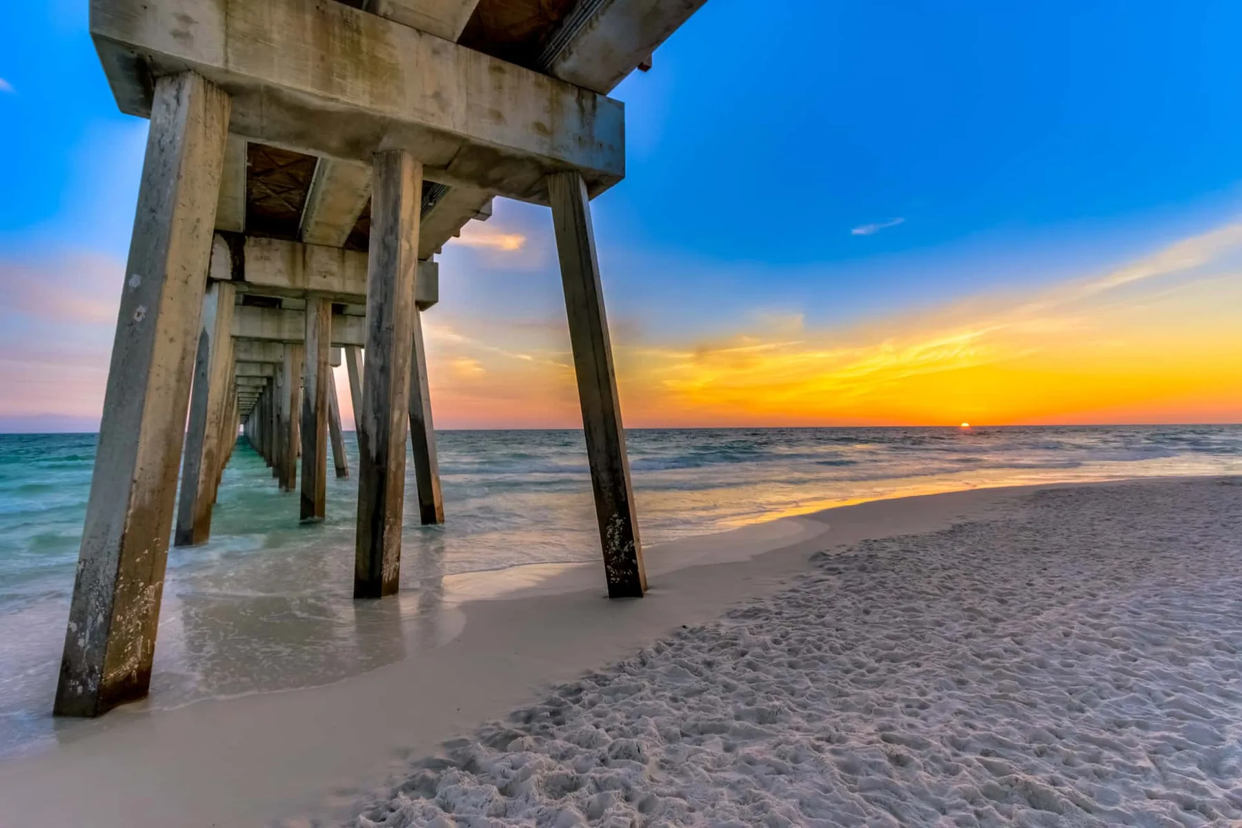 A wooden pier extends over the beach beside calm water at sunset.