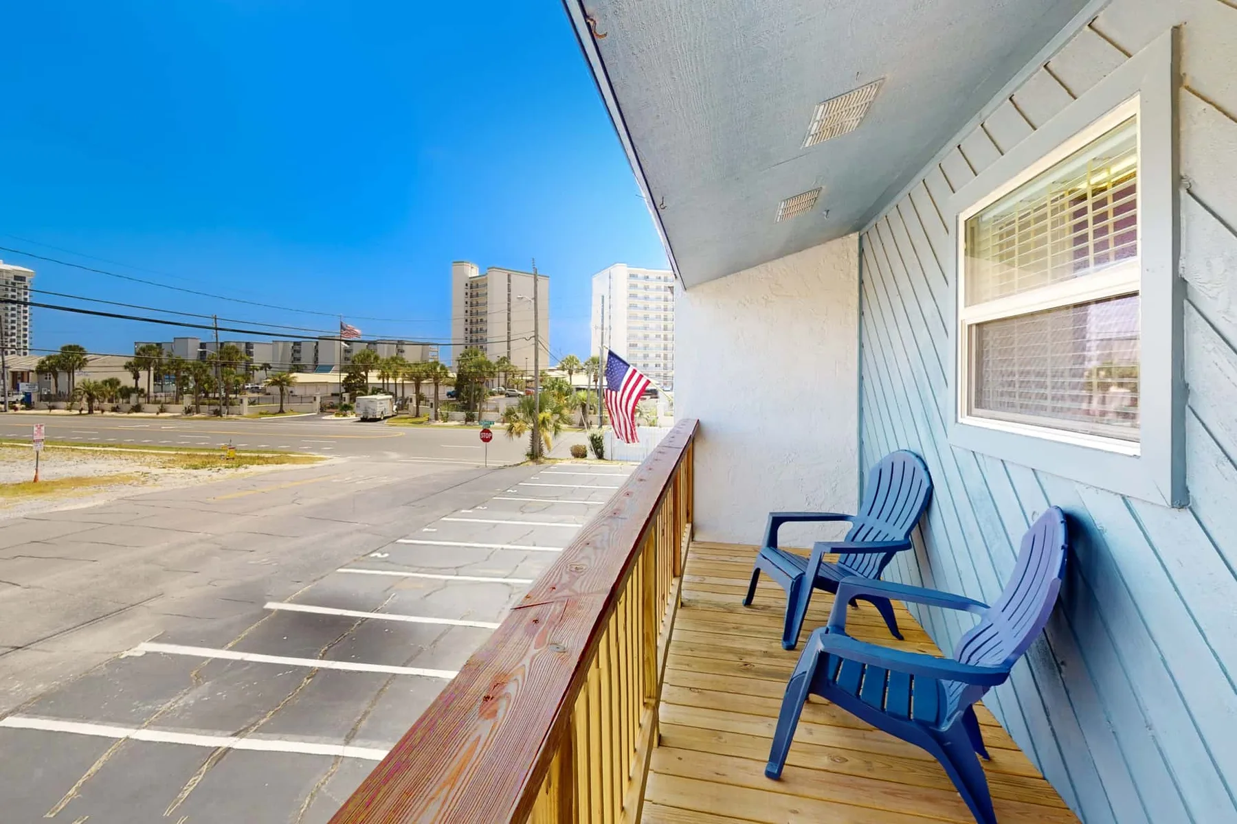 A wooden balcony with blue seating overlooks a parking area and nearby buildings.