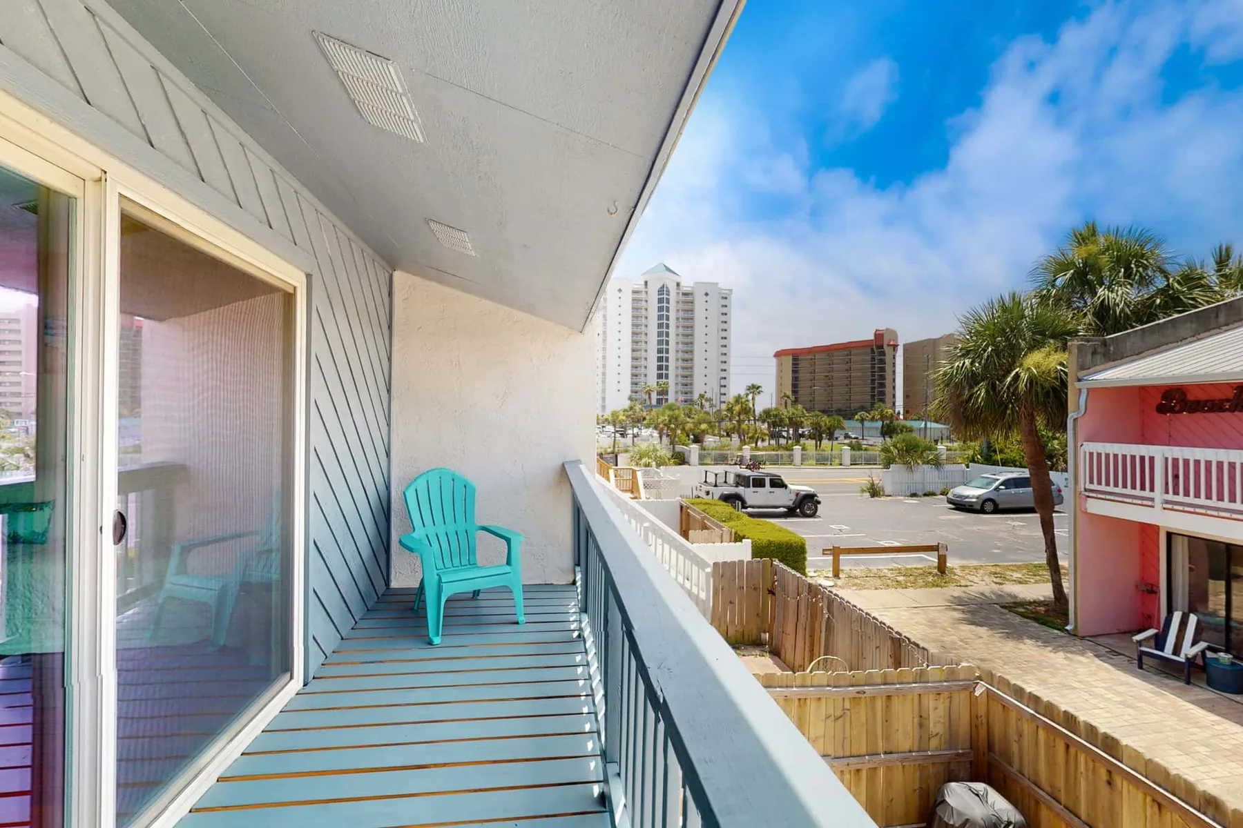 A balcony with a turquoise chair overlooks nearby buildings and a parking area.