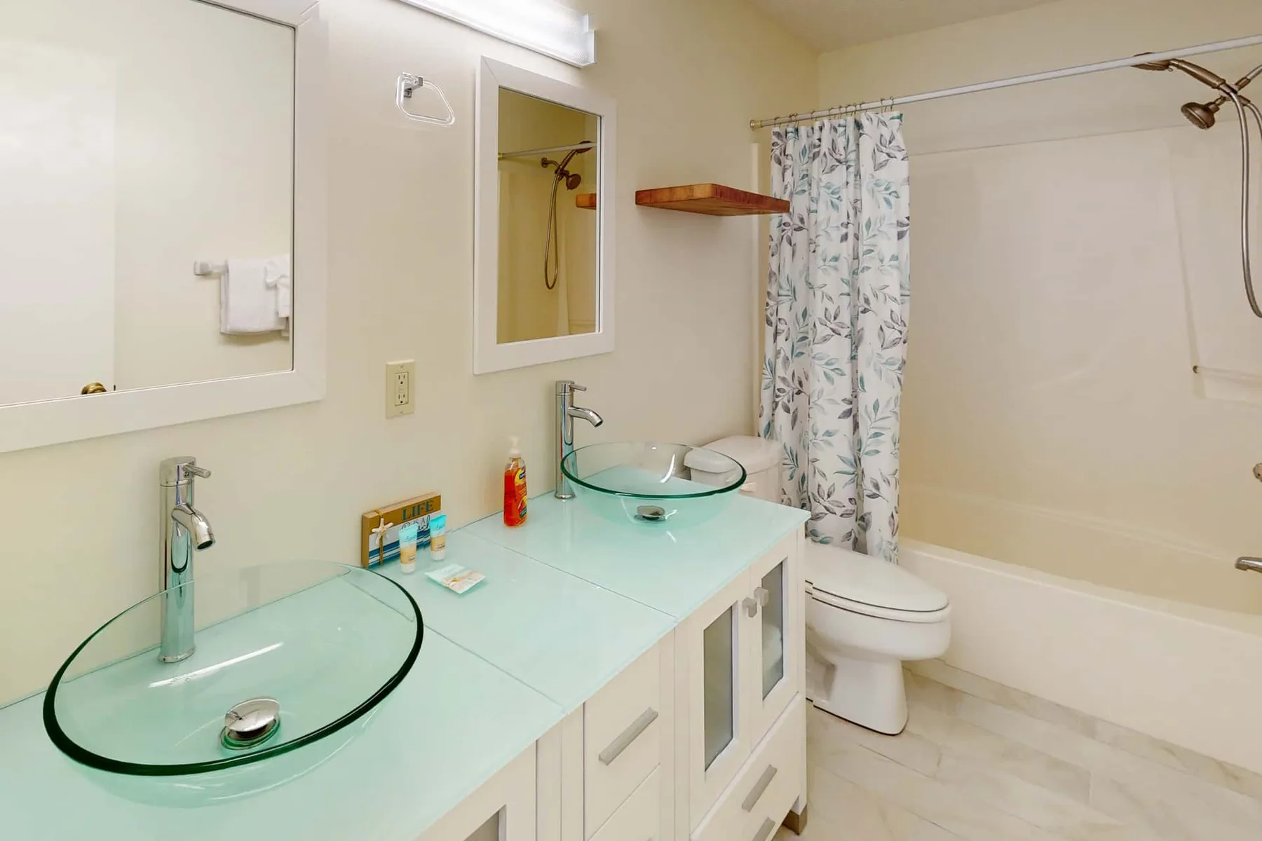 A bathroom with a glass sink, toilet, and bathtub behind a patterned curtain.