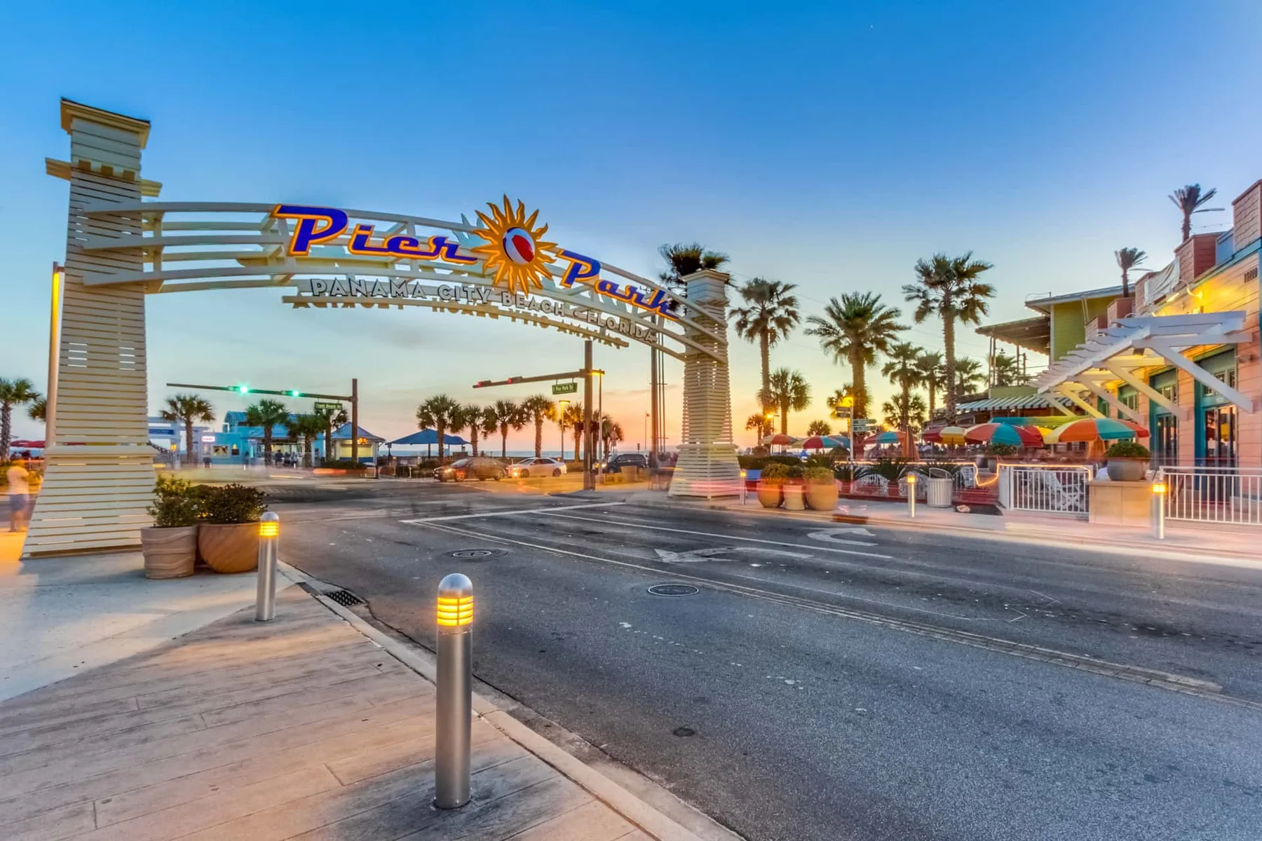 A palm-lined street at sunset passes beneath a colorful overhead sign.