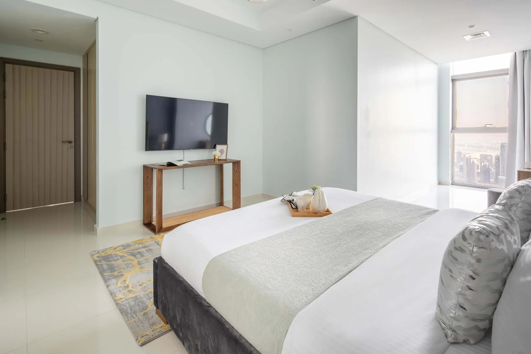 Un dormitorio luminoso con cama, televisor en la pared y una ventana grande.