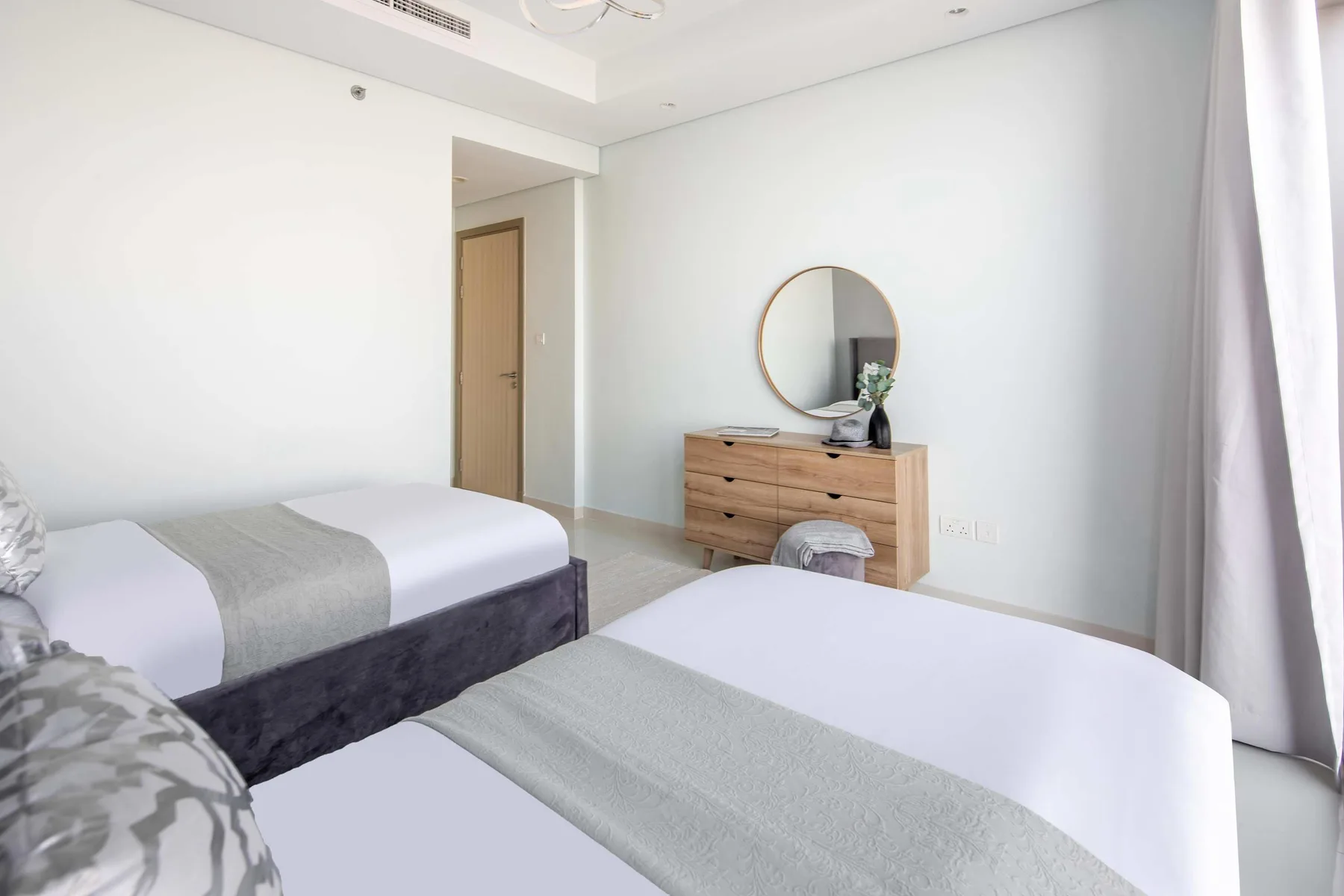 Un dormitorio luminoso con dos camas y una cómoda de madera bajo un espejo redondo.