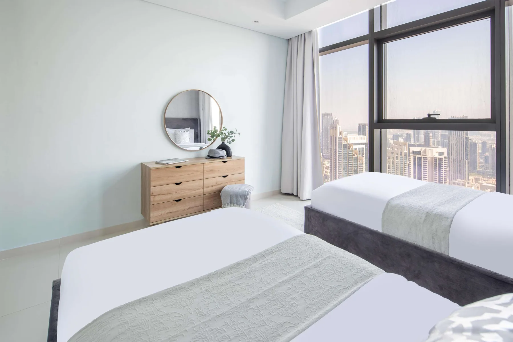 Un dormitorio luminoso con dos camas, una cómoda y grandes ventanas con vistas a edificios de la ciudad.
