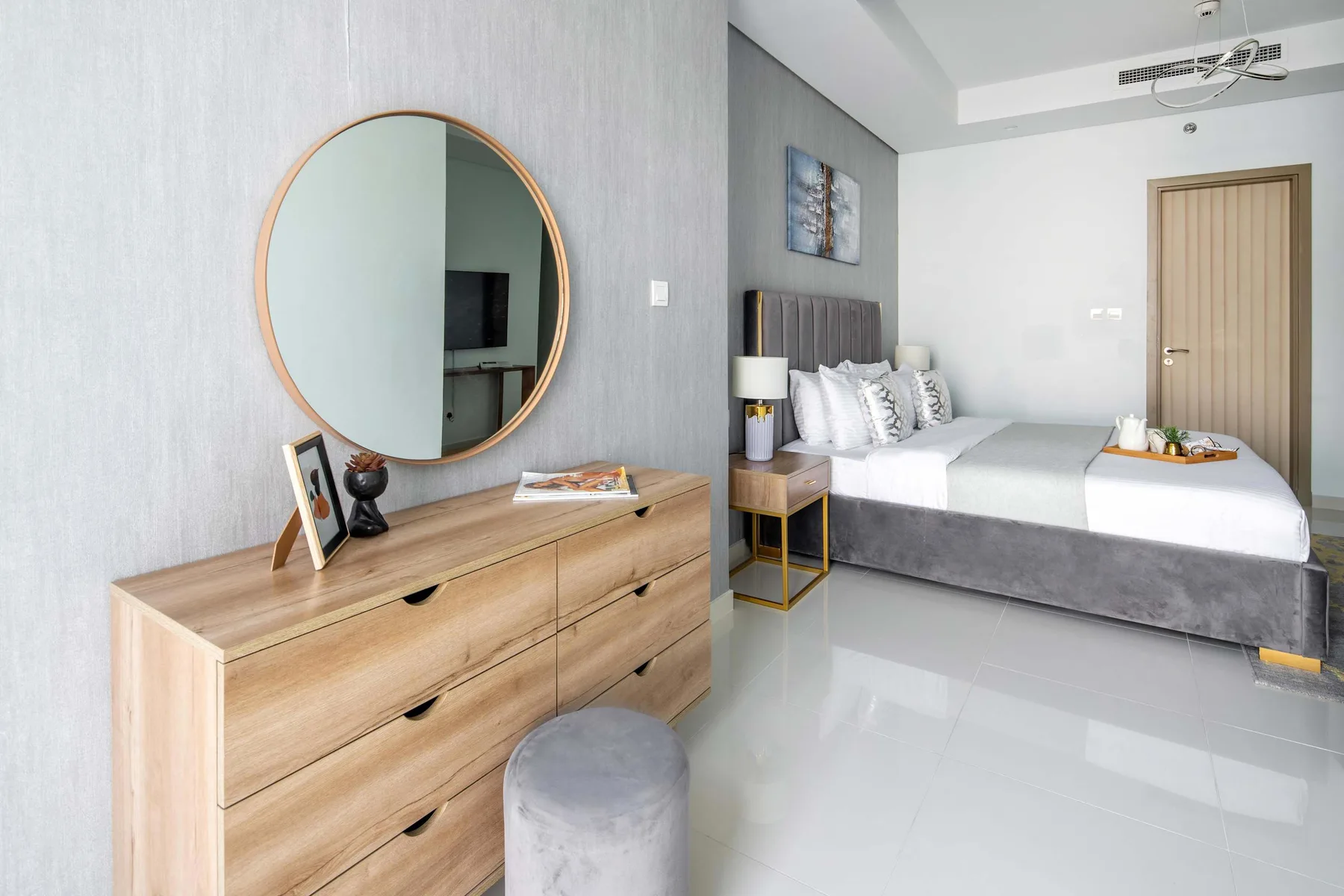 Un dormitorio luminoso con una cama y una cómoda de madera bajo un espejo redondo.