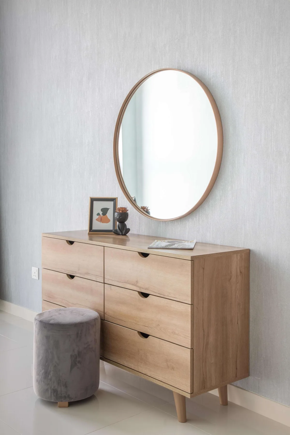 Una cómoda de madera clara está bajo un espejo ovalado, con un pequeño taburete cerca.