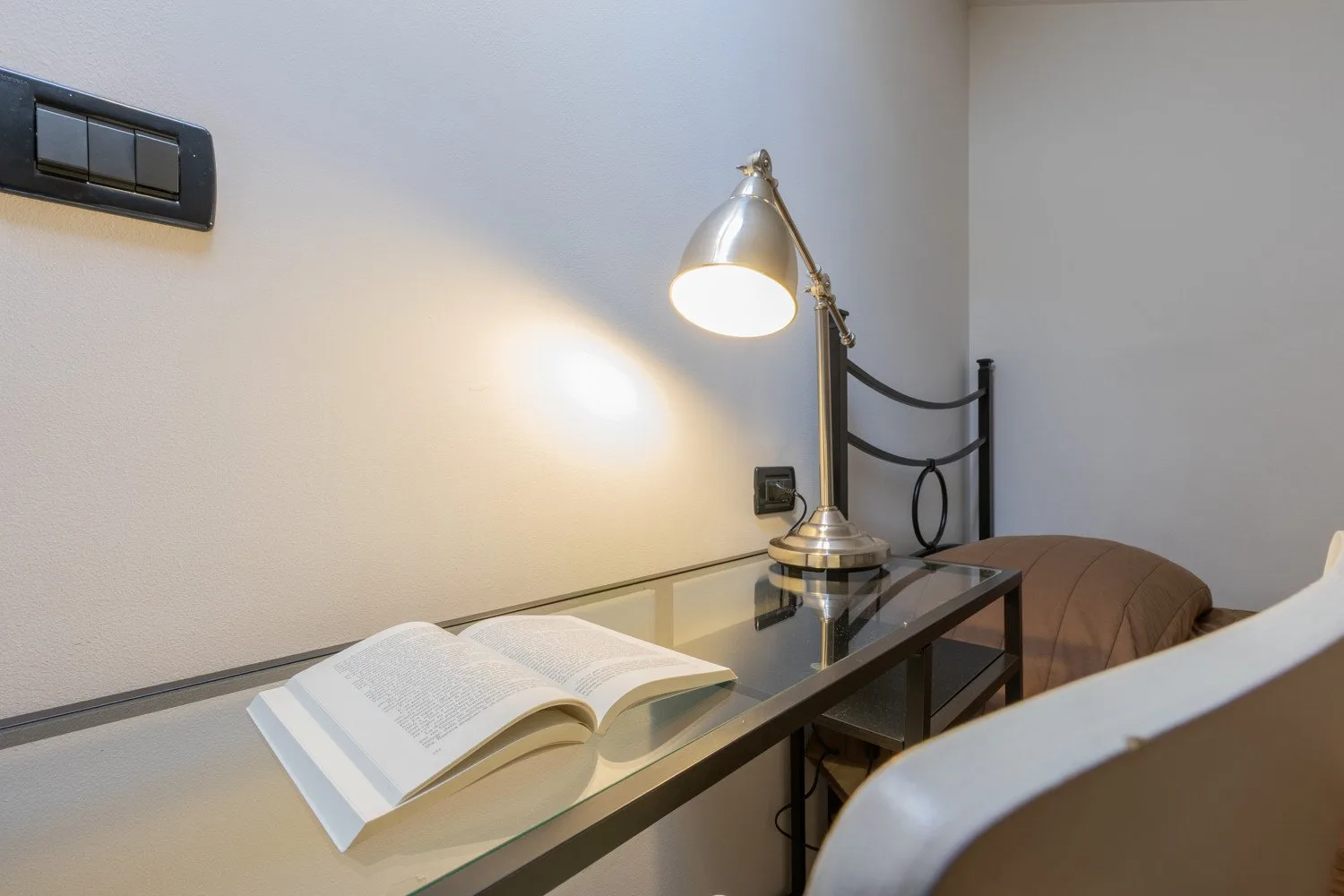 Un espacio de trabajo en un dormitorio, con un libro abierto y una lámpara encendida.