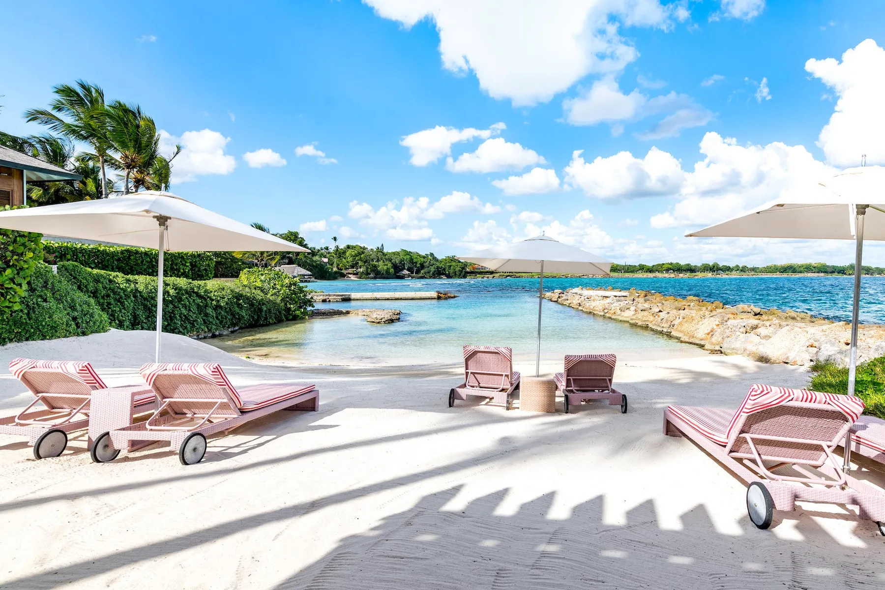 A sunny beachside lounge area has striped seating, umbrellas, and a view of the water.