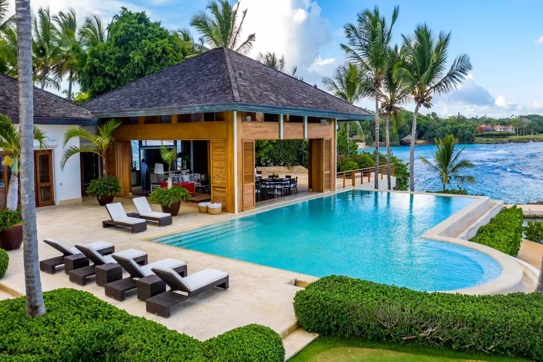 A swimming pool beside an open-sided pavilion, with palm trees and water beyond.