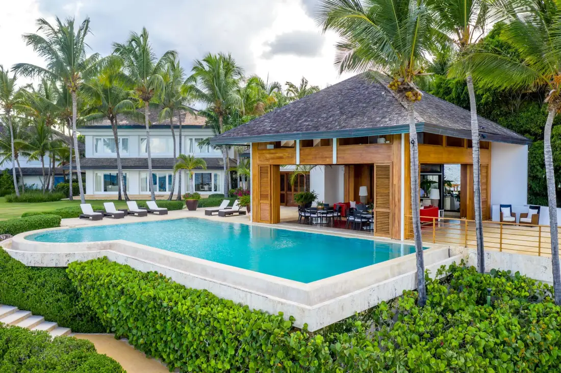 A tropical pool beside a wooden pavilion, with palm trees and a landscaped garden.