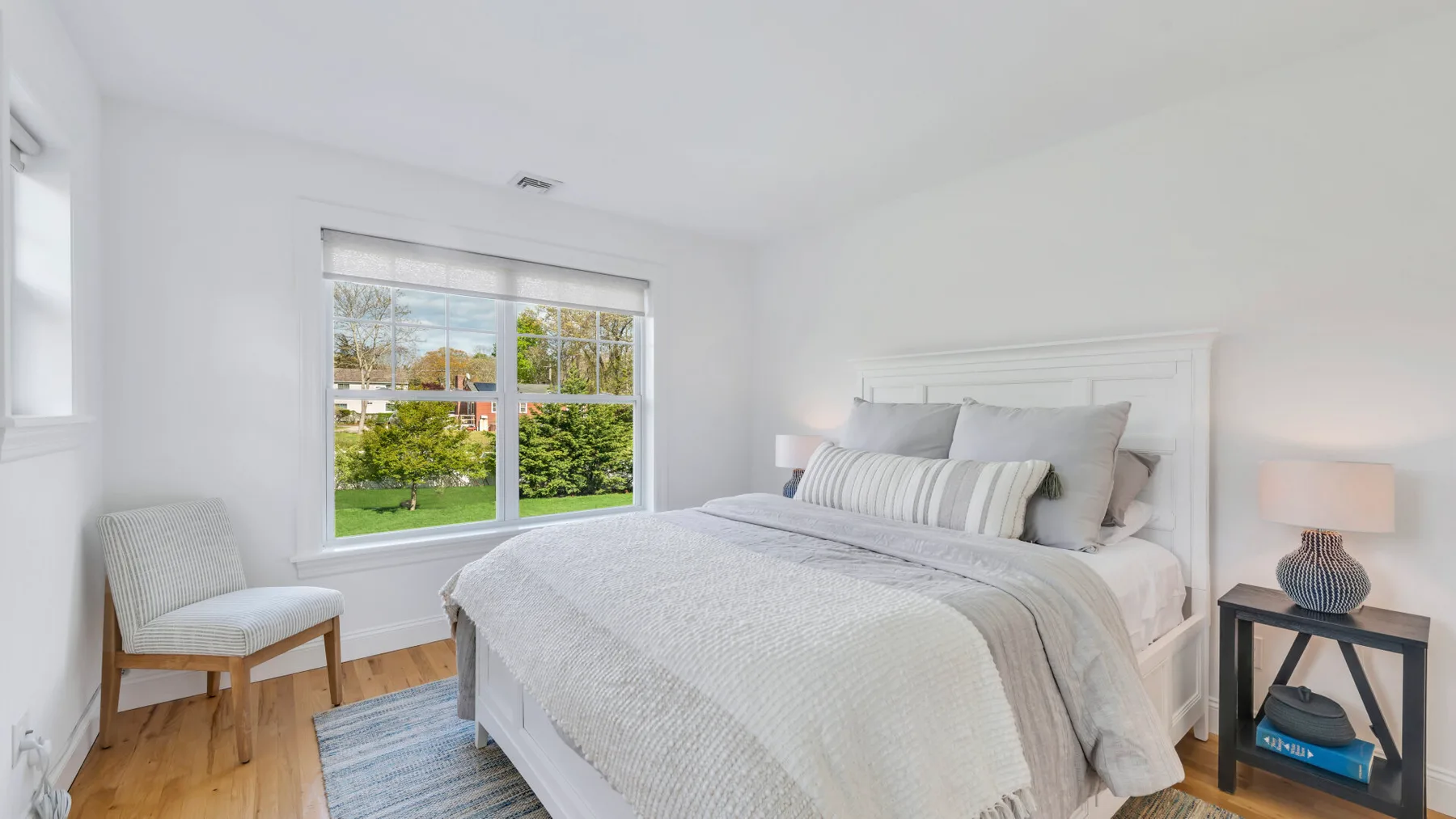 A bright bedroom with a neatly made bed beside a large window overlooking greenery.