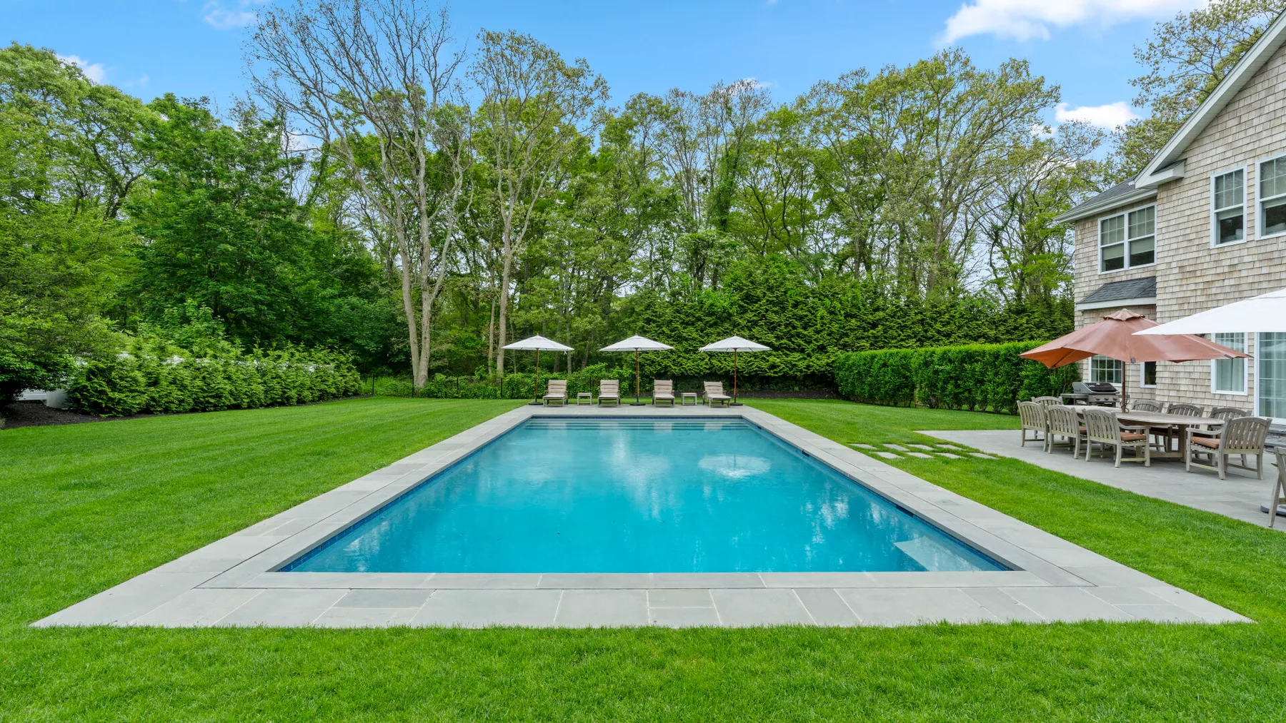 An outdoor swimming pool sits in a landscaped yard beside a house.