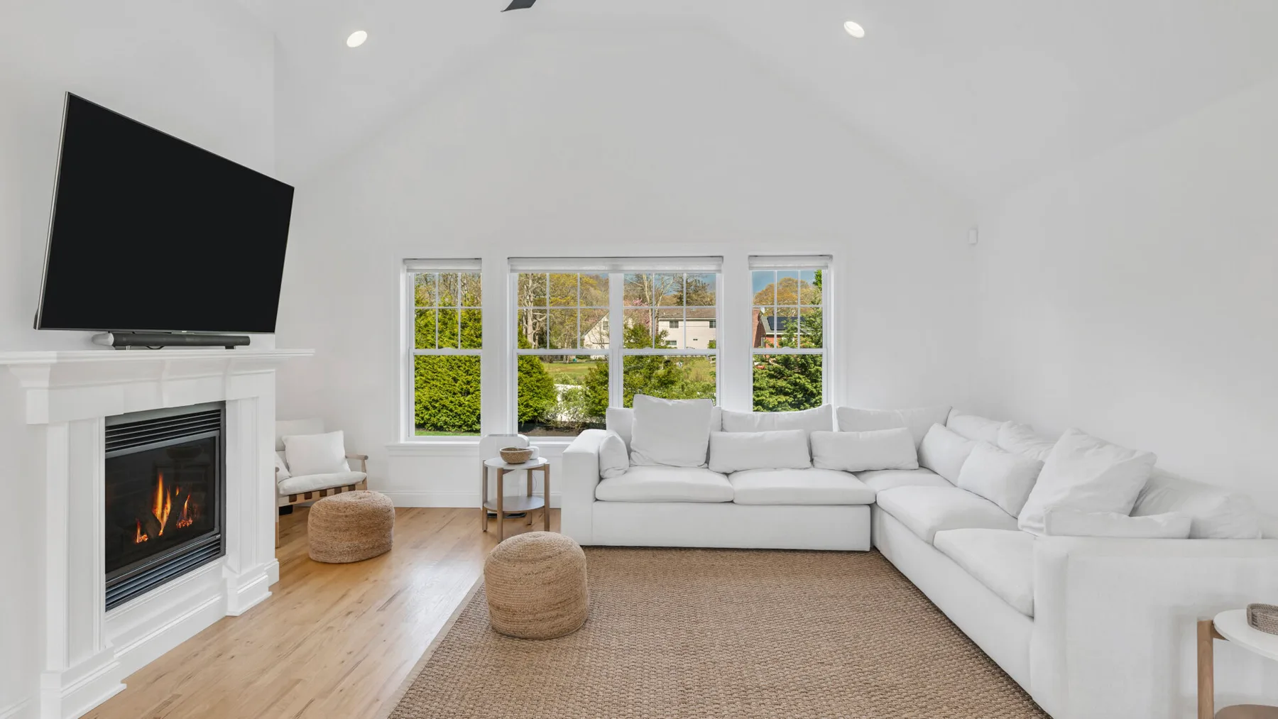 A bright living room with white seating, a fireplace, and windows overlooking greenery.