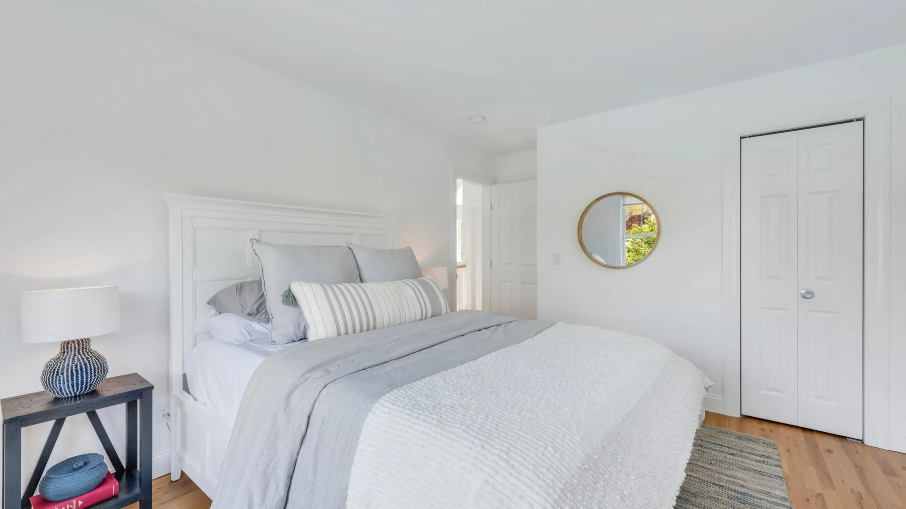 A bright bedroom with a neatly made bed and a round wall mirror.