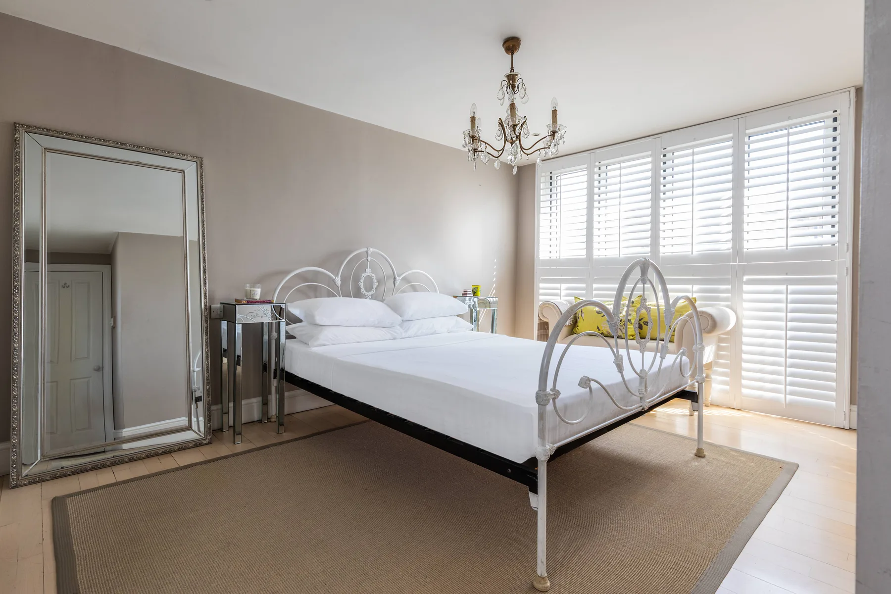 Un dormitorio luminoso con una cama blanca, un espejo grande y ventanas con persianas.