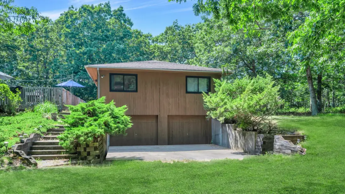 A brown house stands at the edge of a green lawn, surrounded by trees.