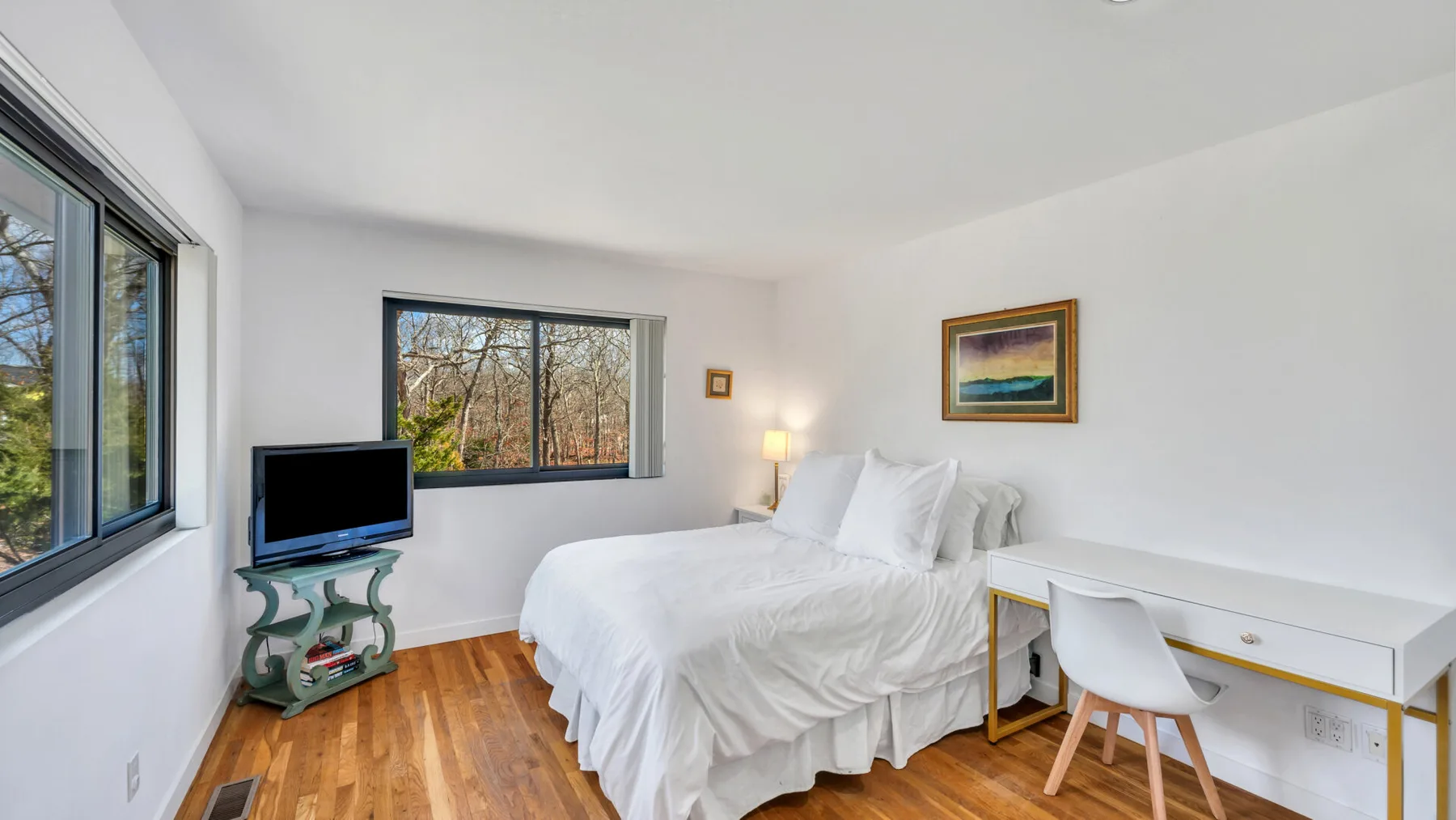 A bright bedroom with a bed, desk, and large windows overlooking trees.