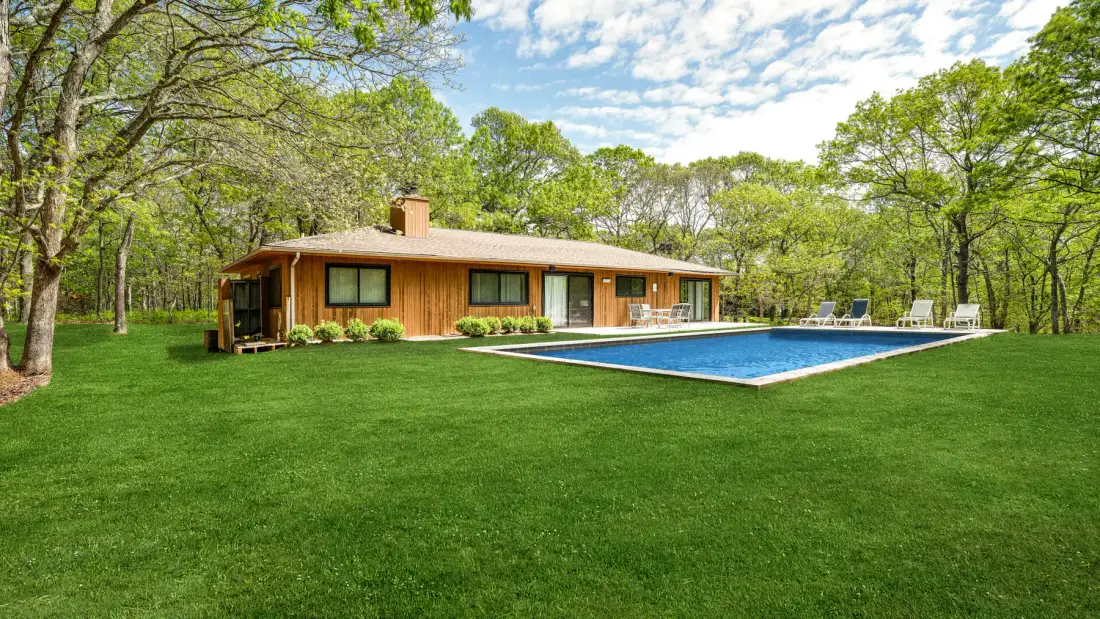Una casa baja de madera está junto a una piscina azul en un amplio césped verde.
