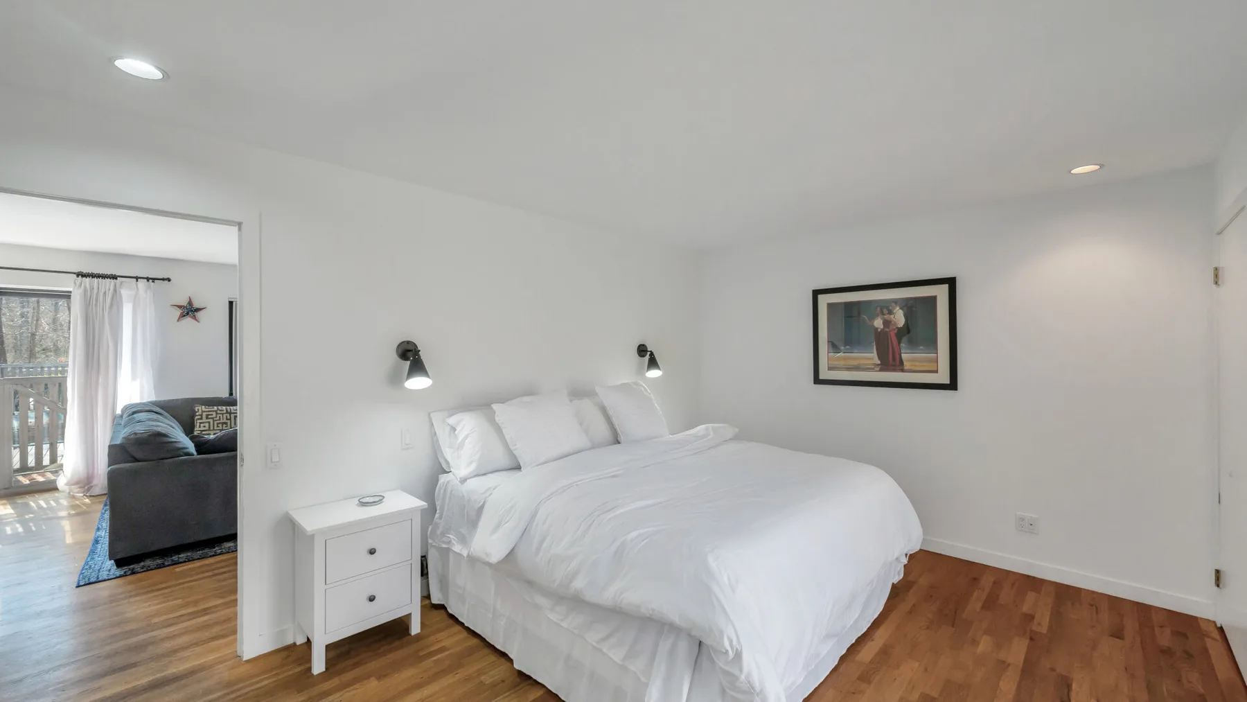 A bright bedroom with a neatly made bed and a doorway to the living area.