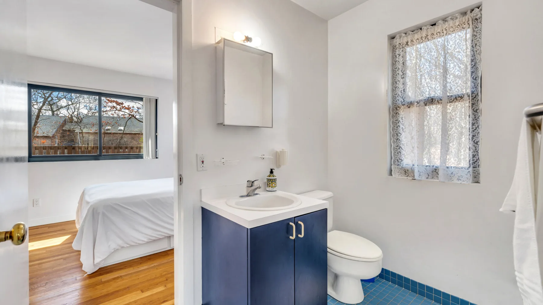 A bathroom with a blue vanity, toilet, and window overlooking a bedroom.