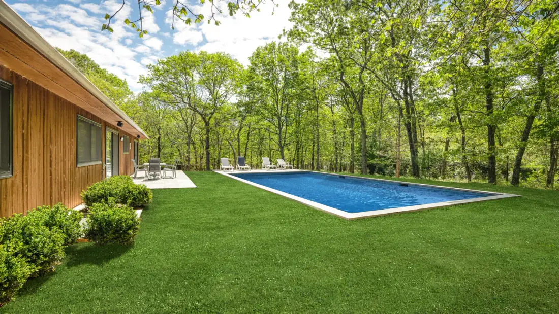 Una piscina y césped verde junto a un edificio revestido de madera, rodeados de árboles.