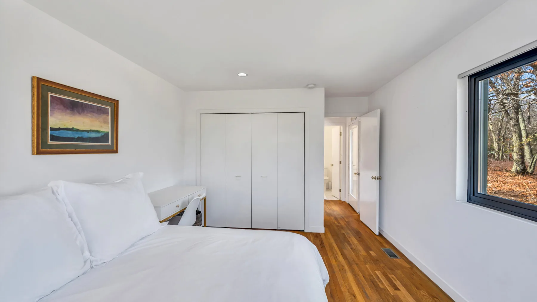 A bright bedroom with a white bed, built-in closets, and a window overlooking trees.
