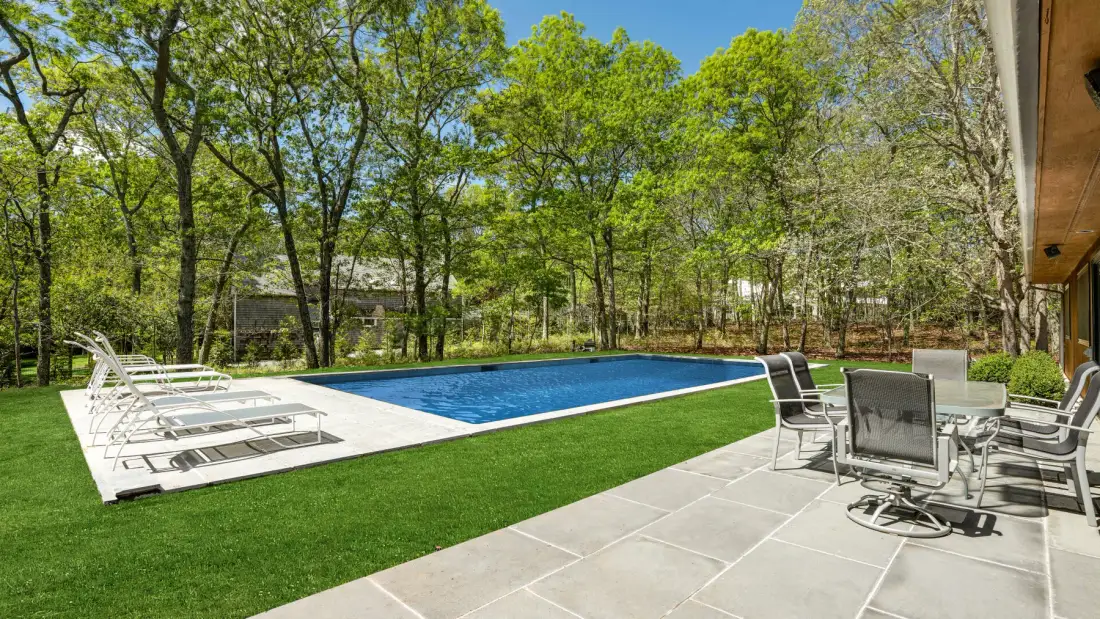 Una piscina al aire libre junto a un patio pavimentado con asientos y árboles alrededor.