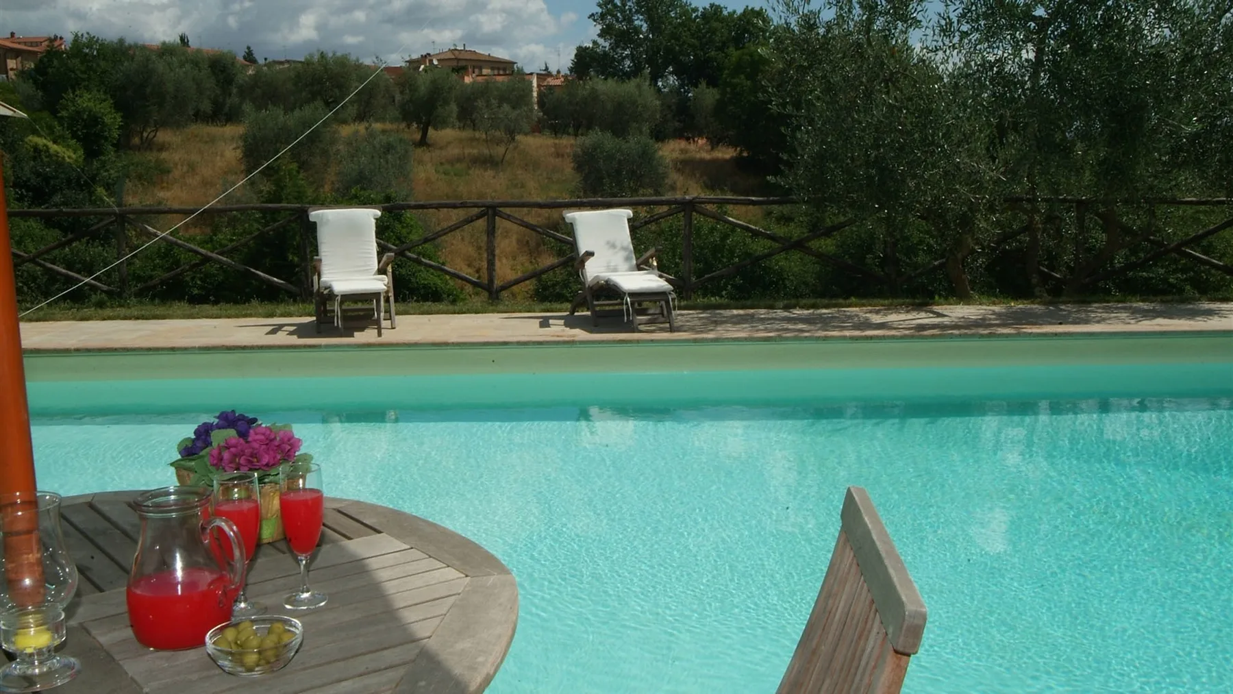 Piscina al aire libre junto a una terraza de madera con bebidas y flores.