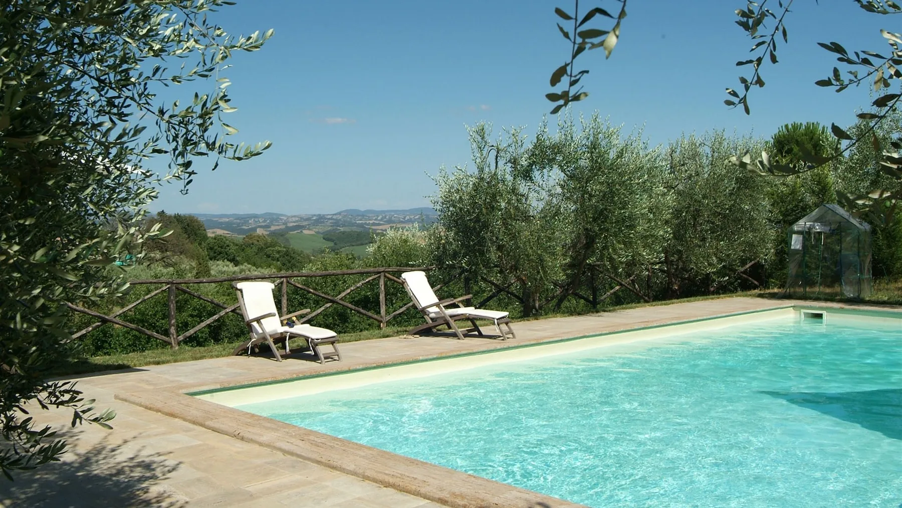Una piscina al aire libre junto a una terraza pavimentada, con árboles y colinas al fondo.