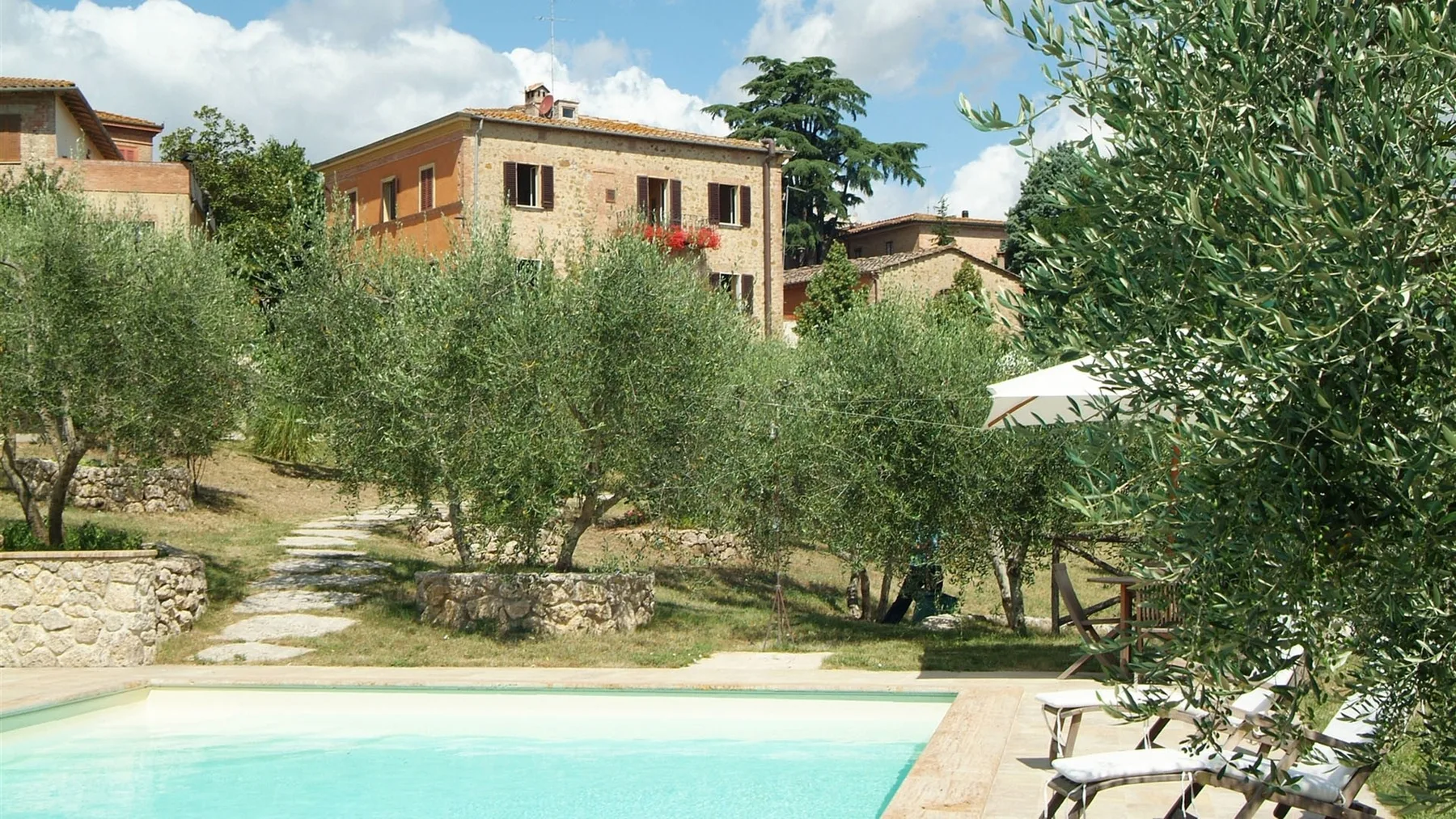 Una piscina al aire libre junto a un jardín con árboles y edificios cercanos.