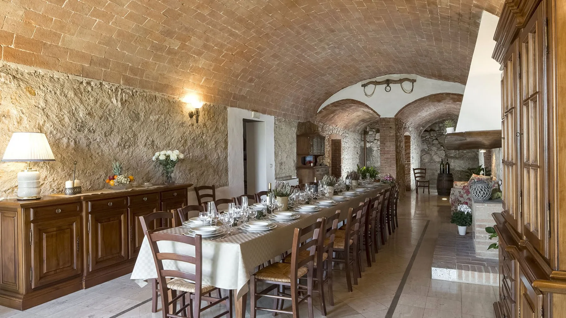 Una larga mesa de comedor está puesta en una rústica sala de piedra con techos abovedados de ladrillo.