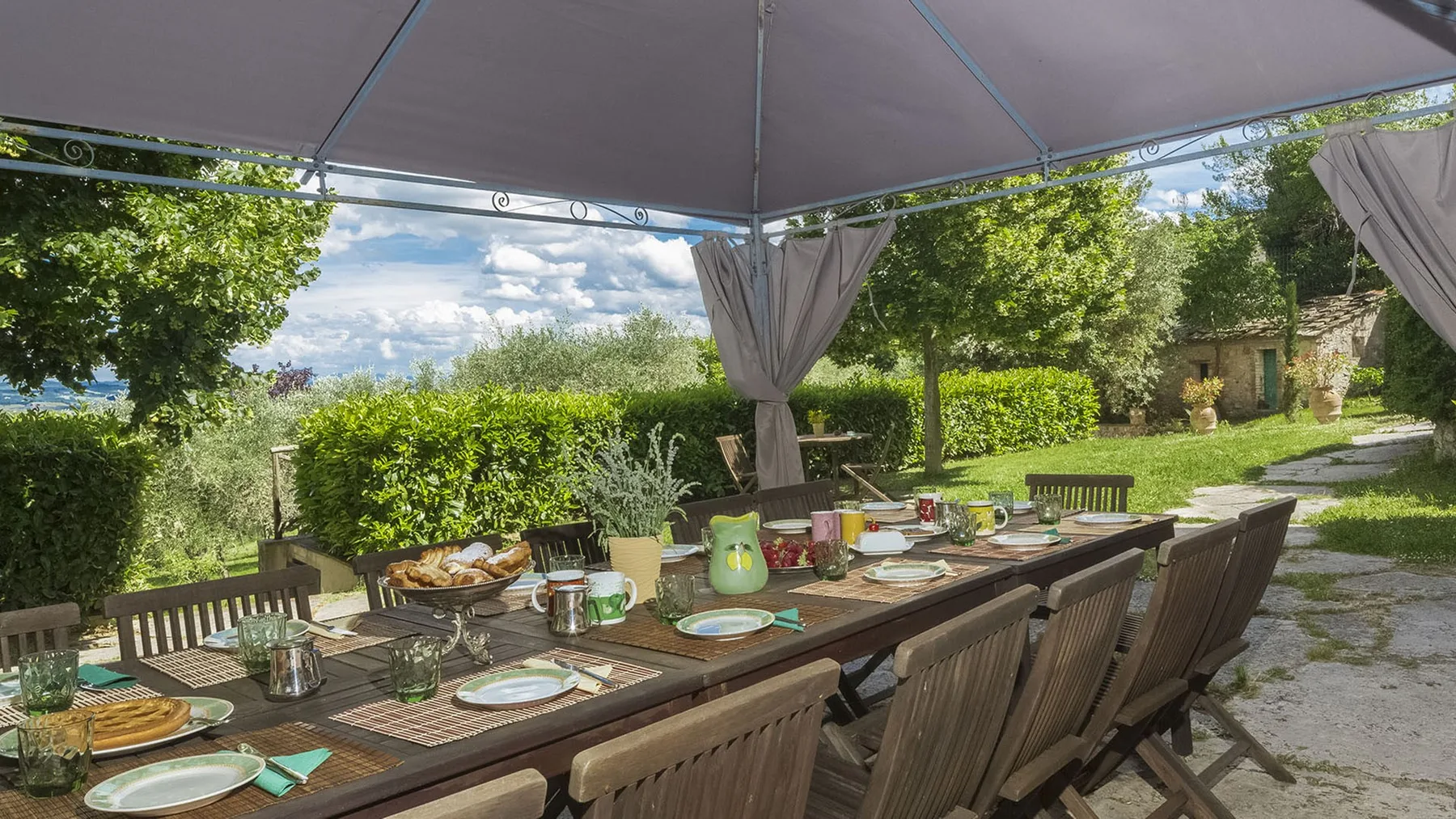 Comedor exterior cubierto con una mesa larga preparada junto a un jardín.