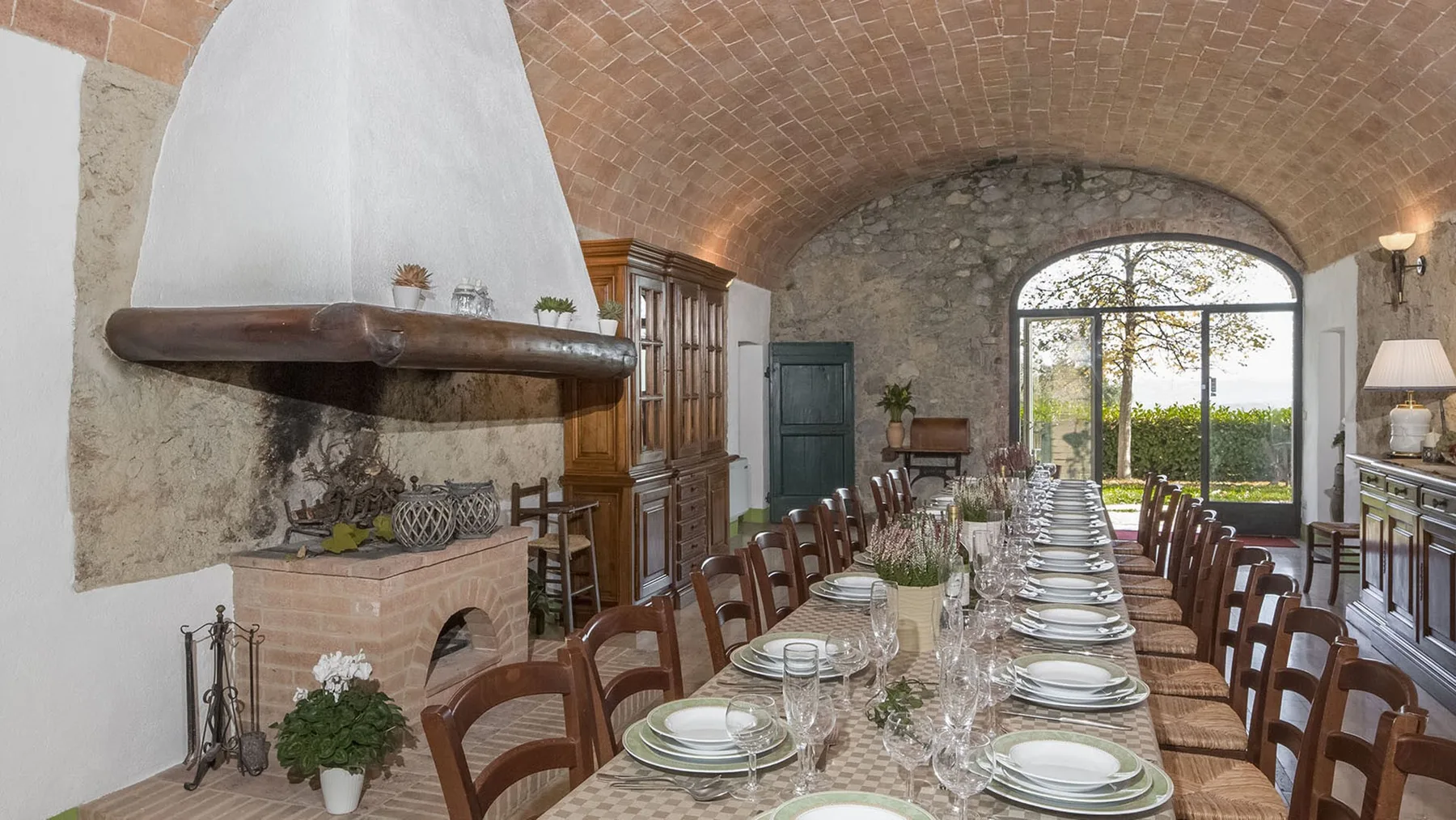 Una larga mesa de comedor está preparada bajo un techo abovedado de ladrillo, junto a una gran ventana arqueada.