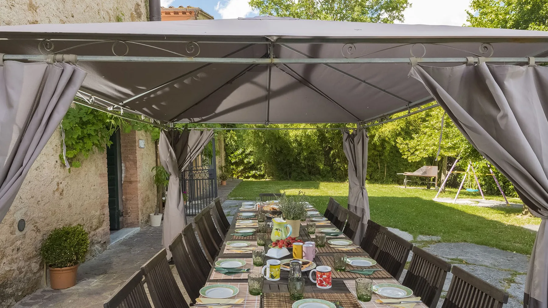 Una zona de comedor exterior cubierta tiene una mesa larga preparada con platos y bebidas.