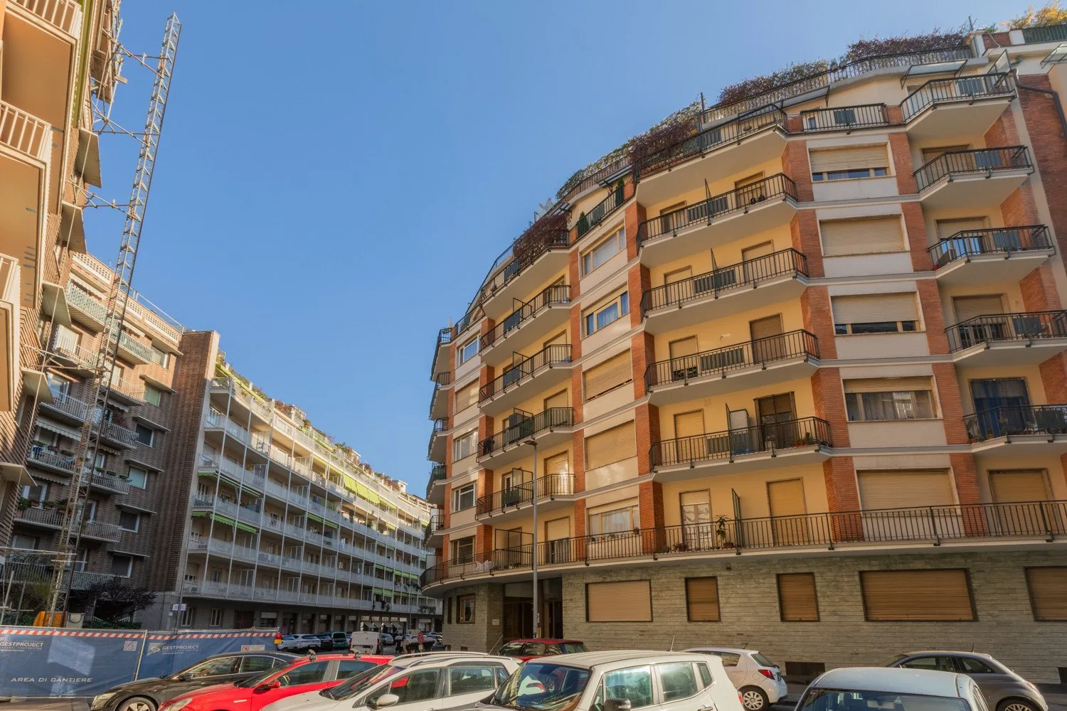 Apartment buildings line an urban street beneath a clear blue sky, with parked cars.