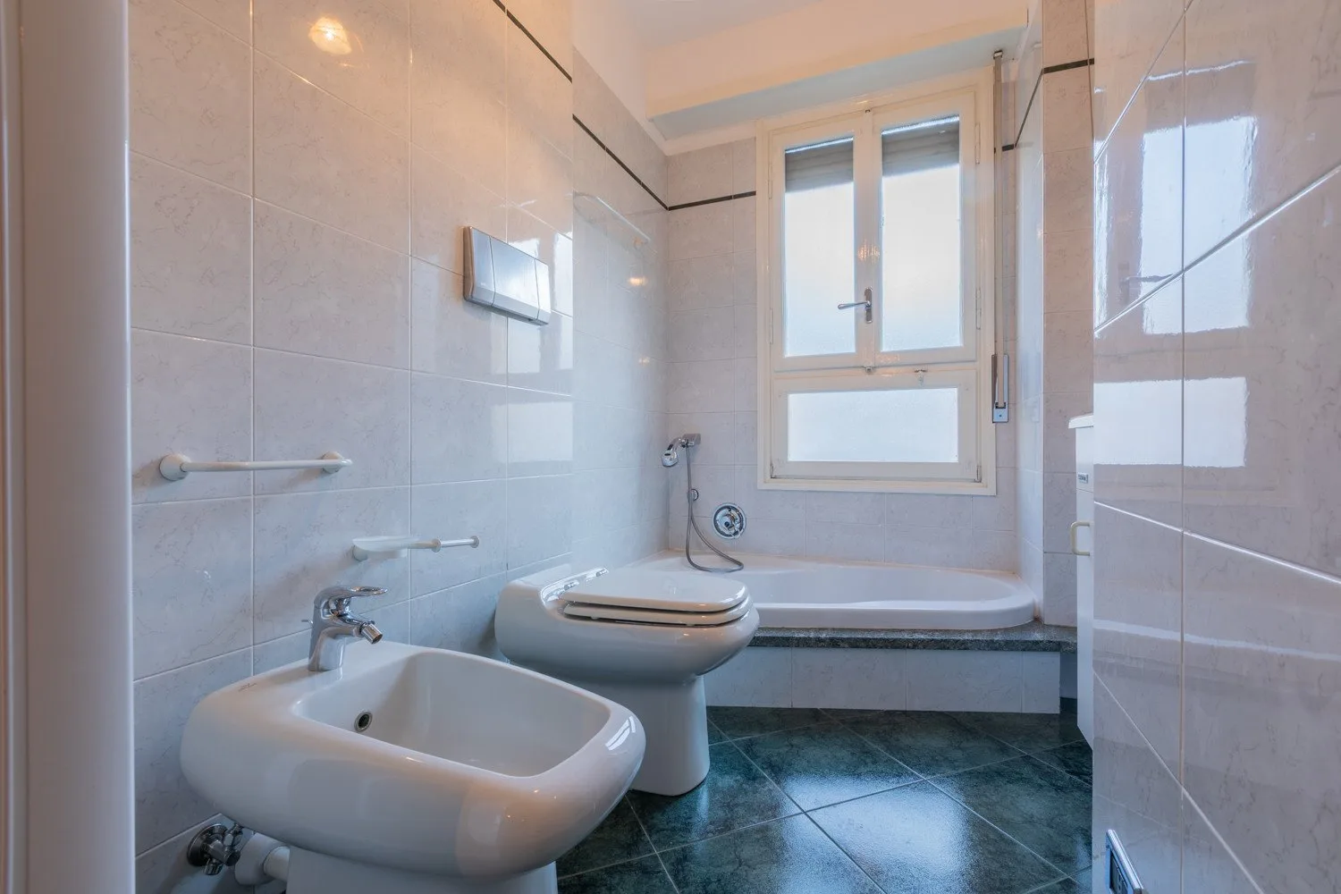 A tiled bathroom with a bathtub, toilet, and bidet beneath a window.