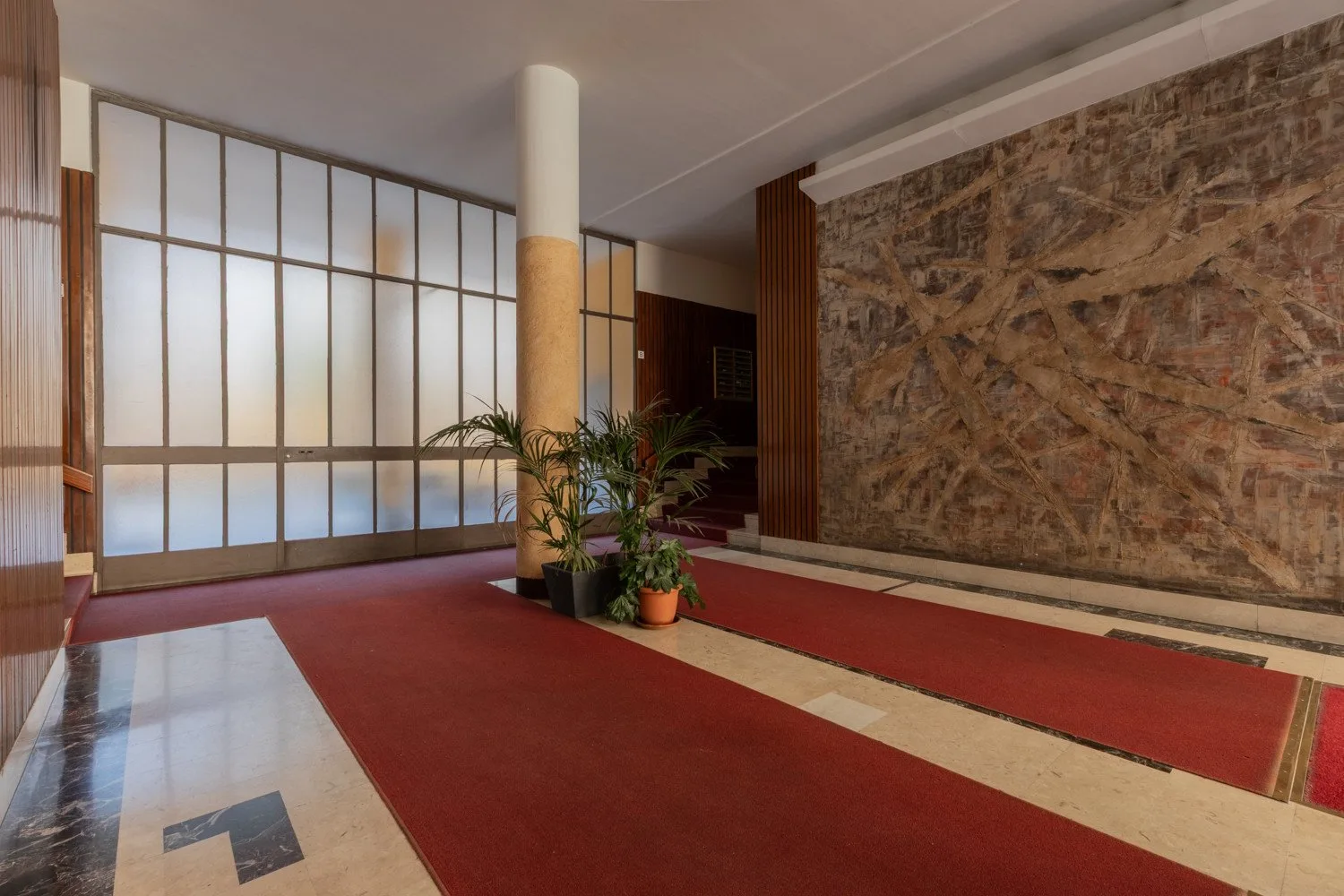 A spacious lobby with red carpet and potted plants beside a large window.