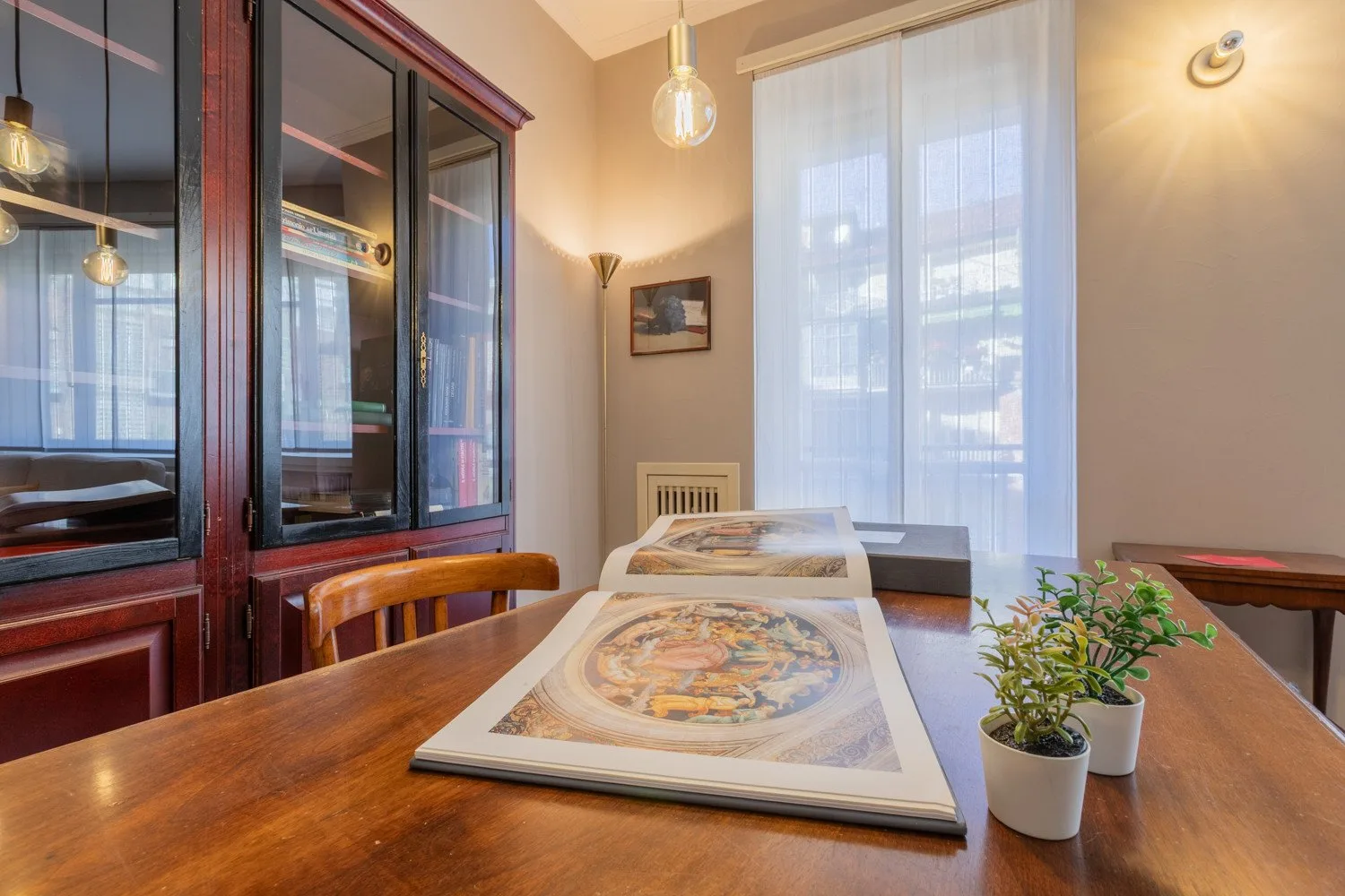 A dining area with a wooden table, framed artwork, and tall curtained windows.