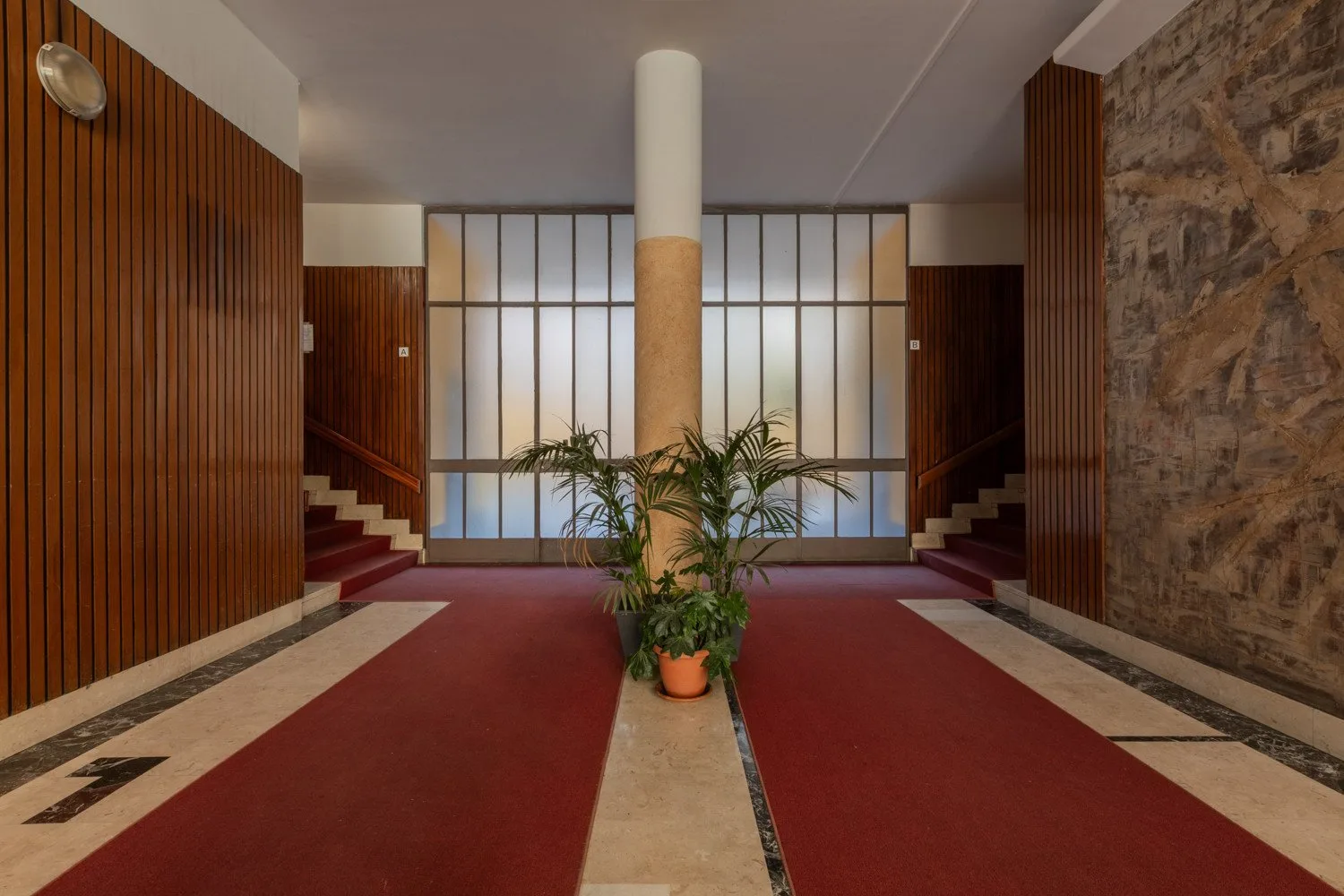A building lobby has red carpet runners, a central column, and potted plants.