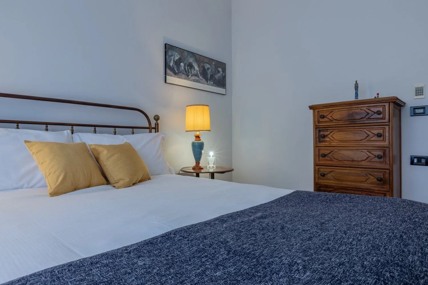 A bedroom with a neatly made bed, bedside lamp, and wooden dresser.