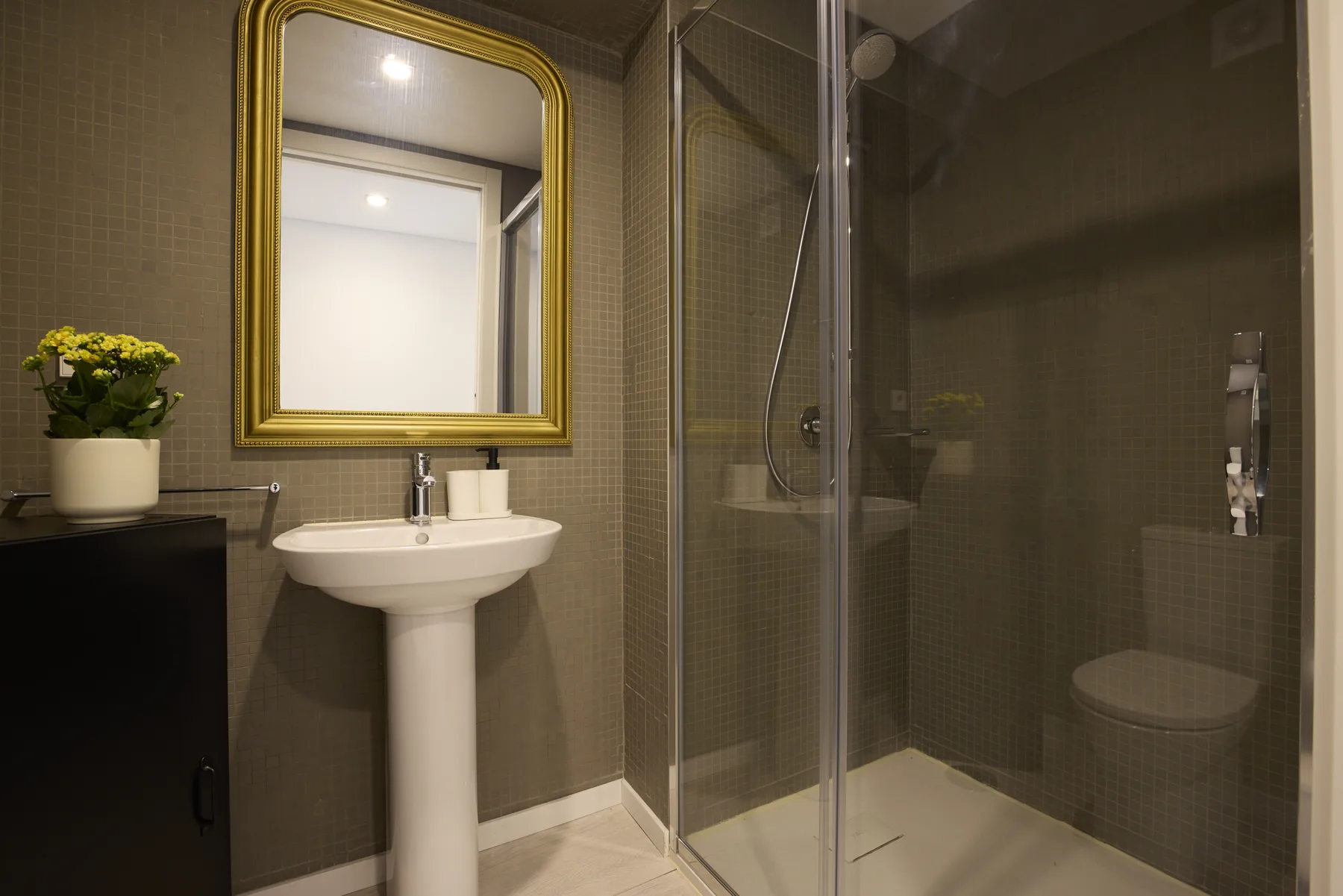 A bathroom with a pedestal sink, gold-framed mirror, and glass shower.