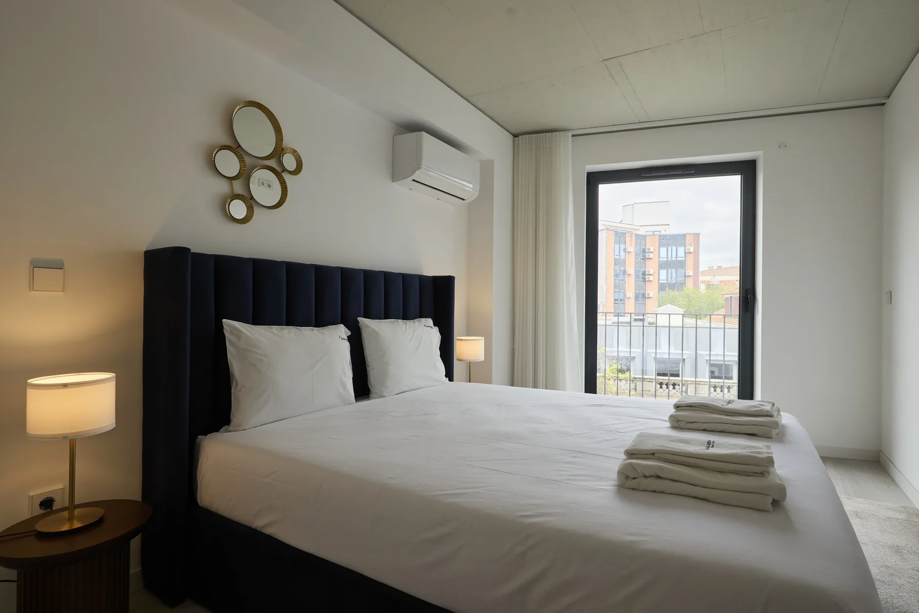 A bedroom with a large bed, bedside lamps, and a window overlooking nearby buildings.