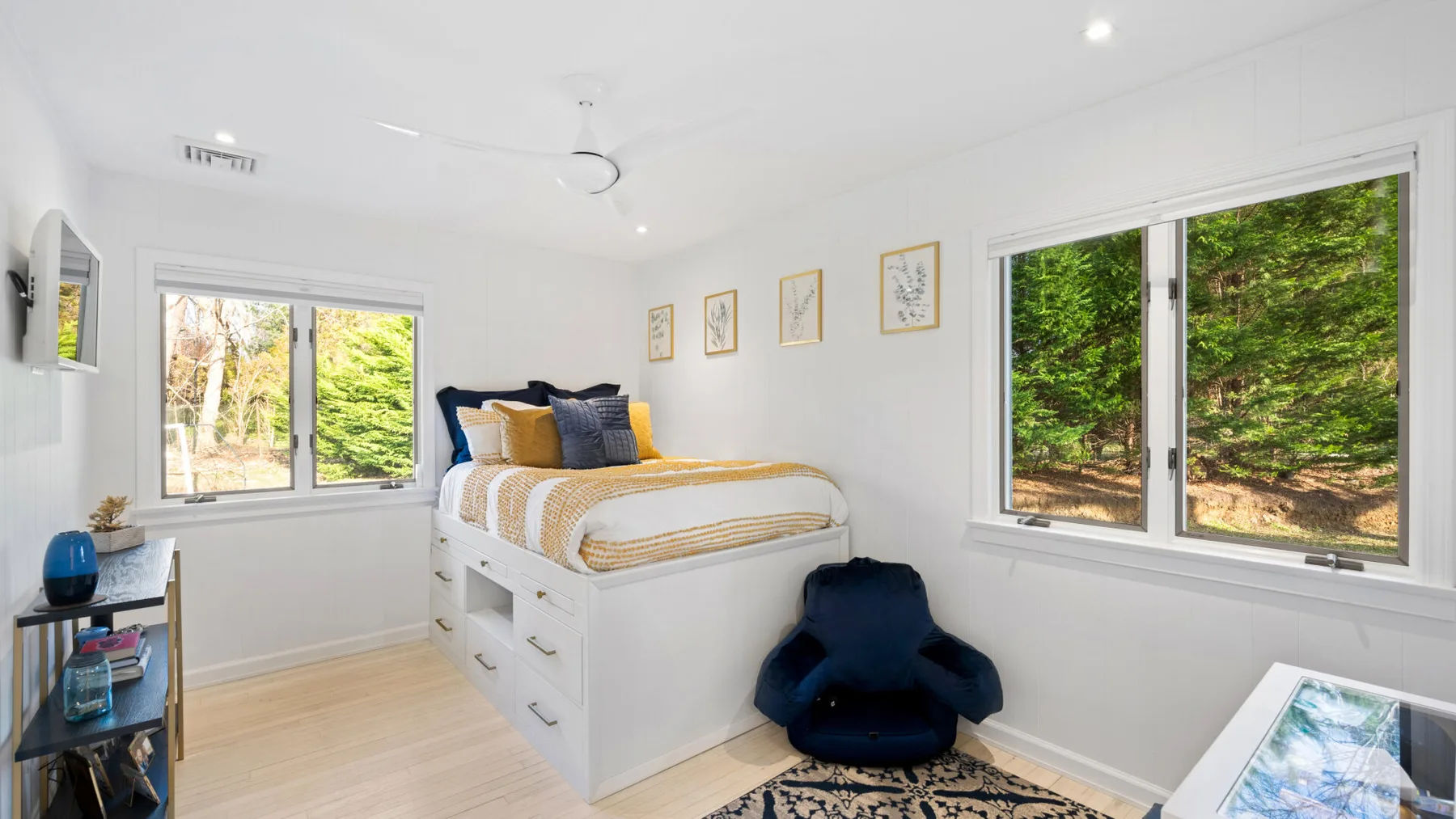 Un dormitorio luminoso tiene una cama elevada con cajones y ventanas con vistas a la vegetación.