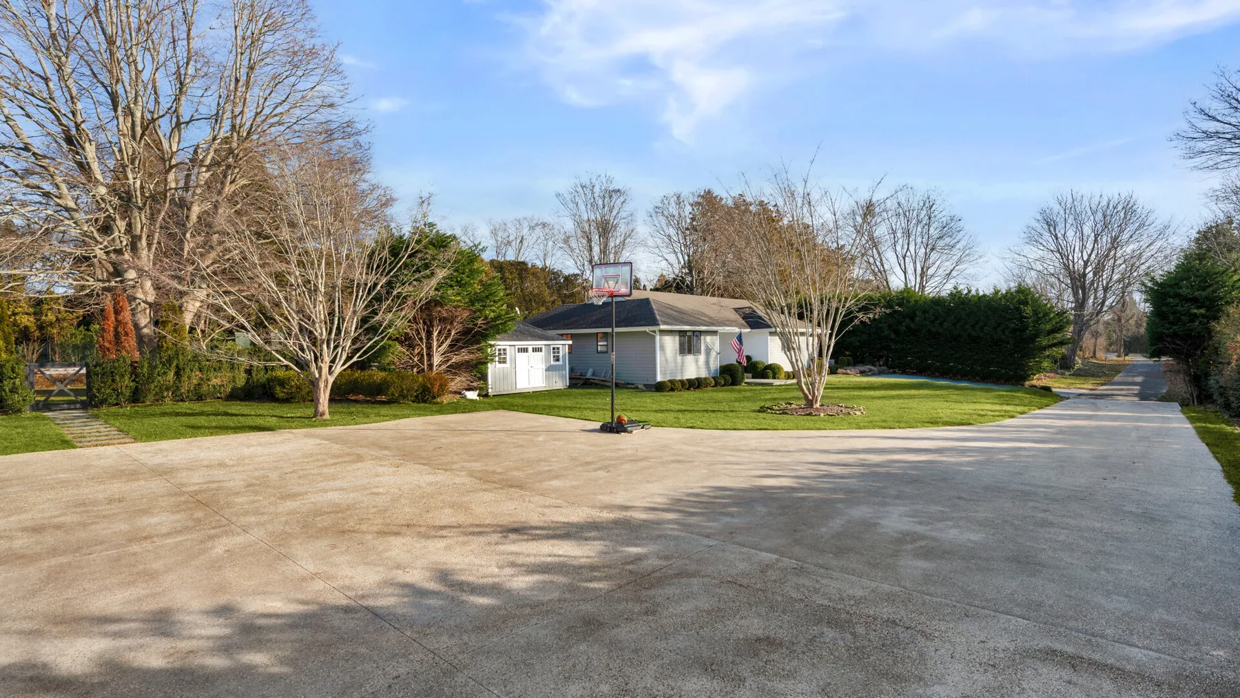 Una cancha exterior pavimentada con una canasta de baloncesto, césped y árboles alrededor.