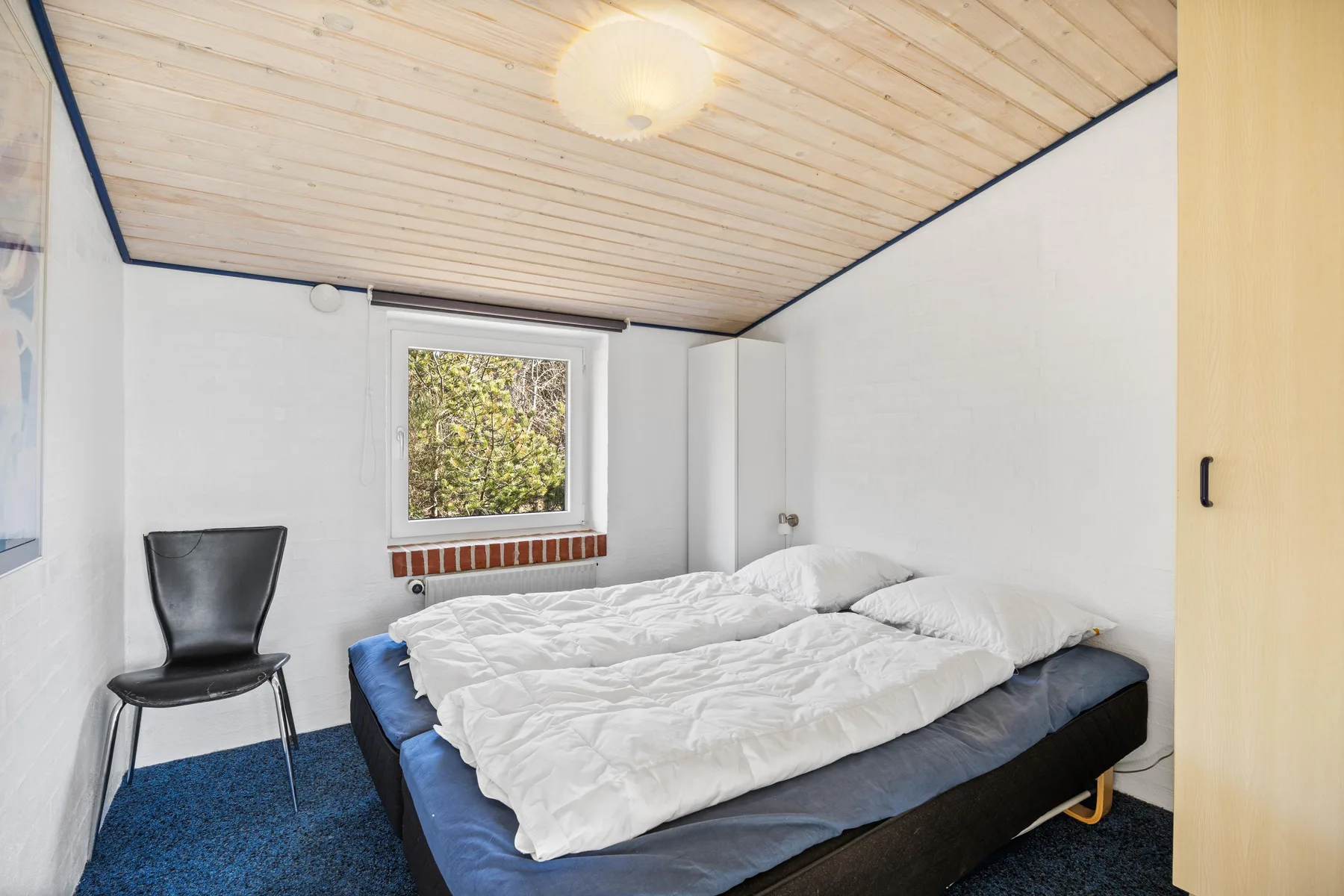 Un dormitorio con una cama bajo una ventana y un techo claro revestido de madera.