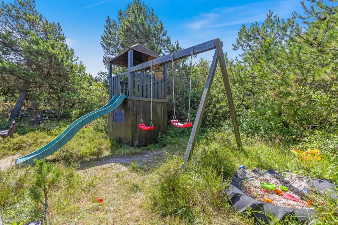 Una estructura de juego al aire libre con tobogán y columpios está entre árboles y hierba.