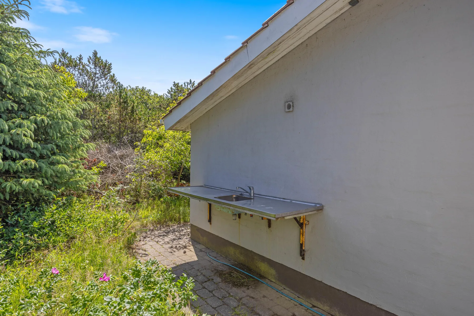 Un patio exterior junto a una pared blanca, con vegetación al lado del sendero pavimentado.