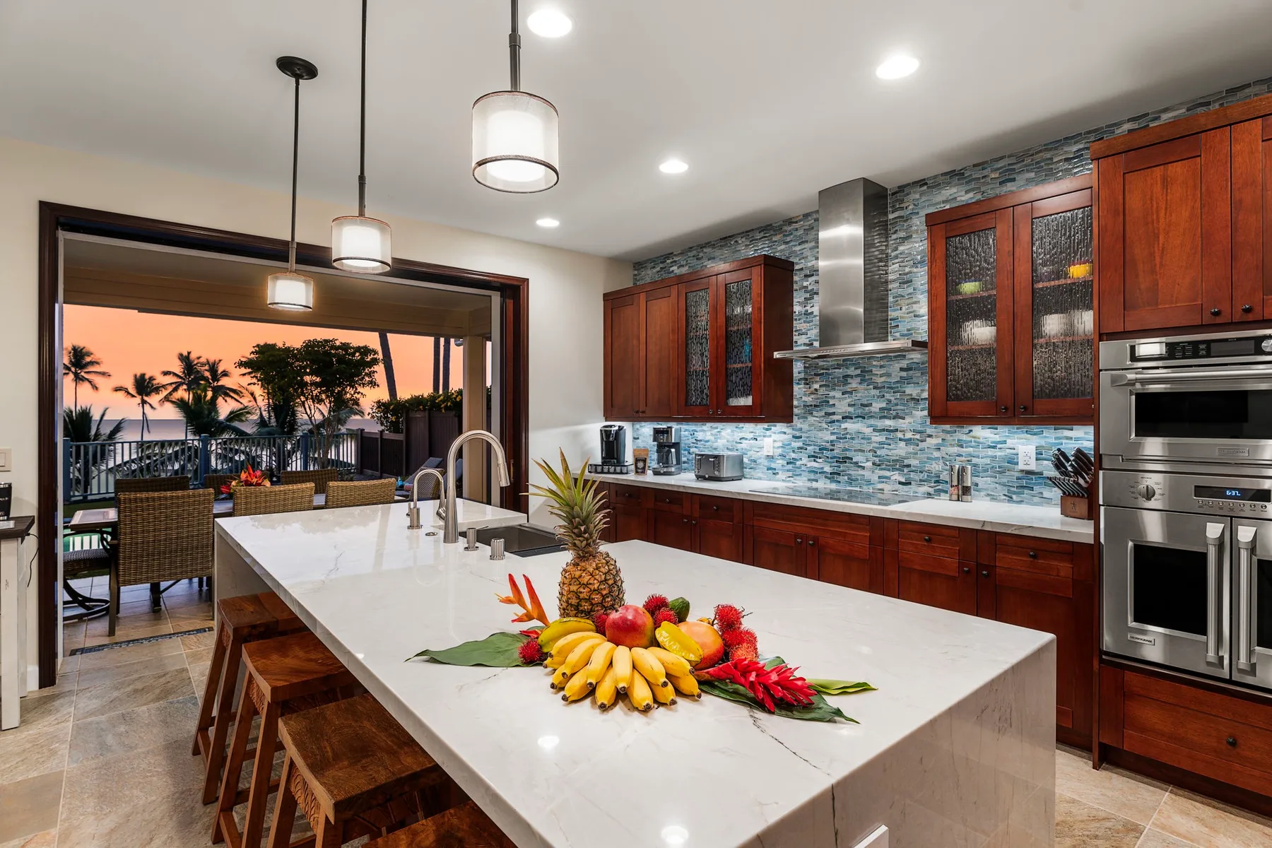 A modern kitchen with a large island, wooden cabinetry, and a sunset view through open doors.