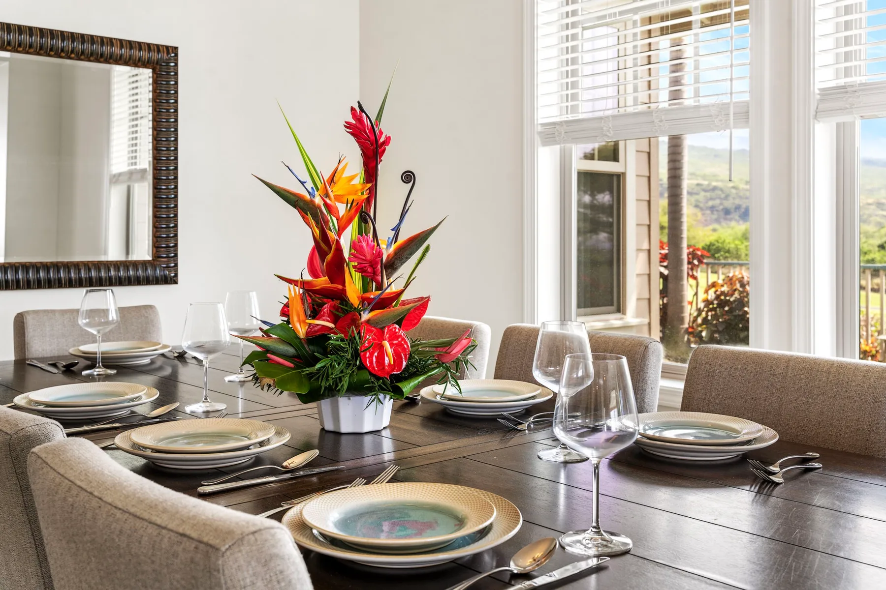 A dining area with a set table and a colorful floral centerpiece beside bright windows.