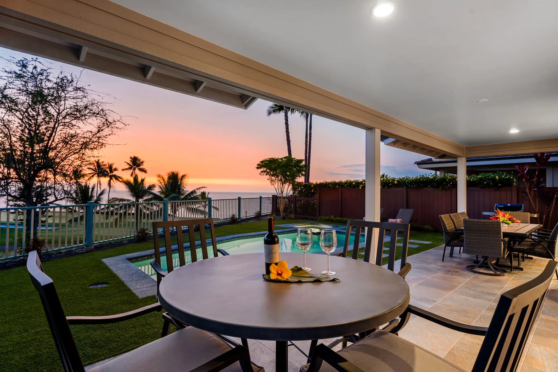 A covered patio dining area overlooks a garden and the ocean at sunset.